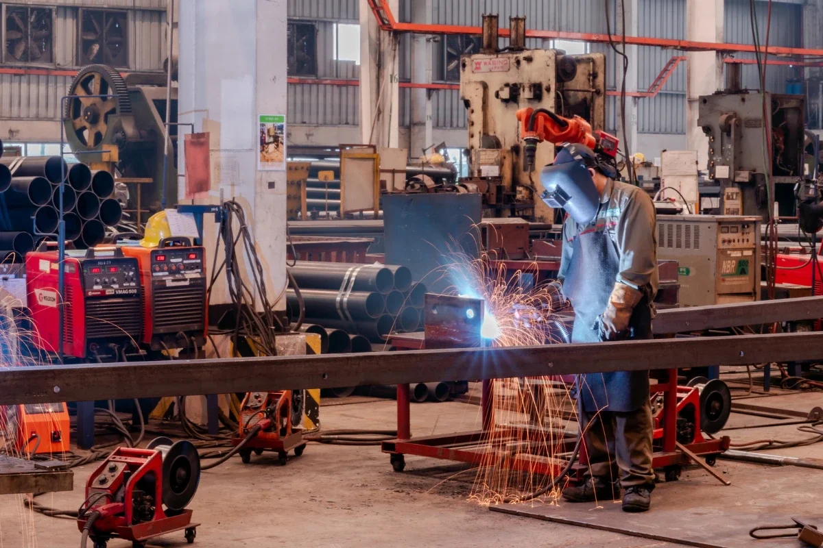 Worker welding metal pipes inside an industrial workshop.