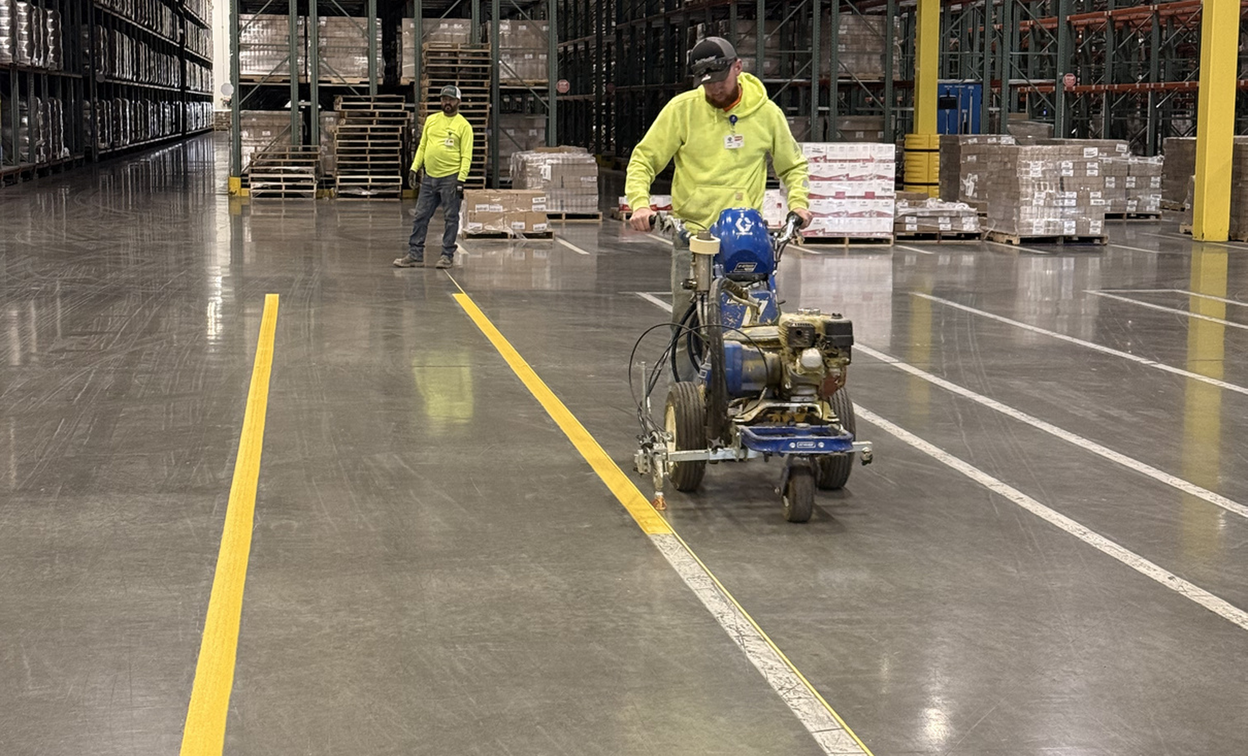 Two workers in high-visibility clothing are working in an industrial warehouse. One worker is operating a large floor grinding or polishing machine, while the other is standing near stacks of pallets filled with boxes.