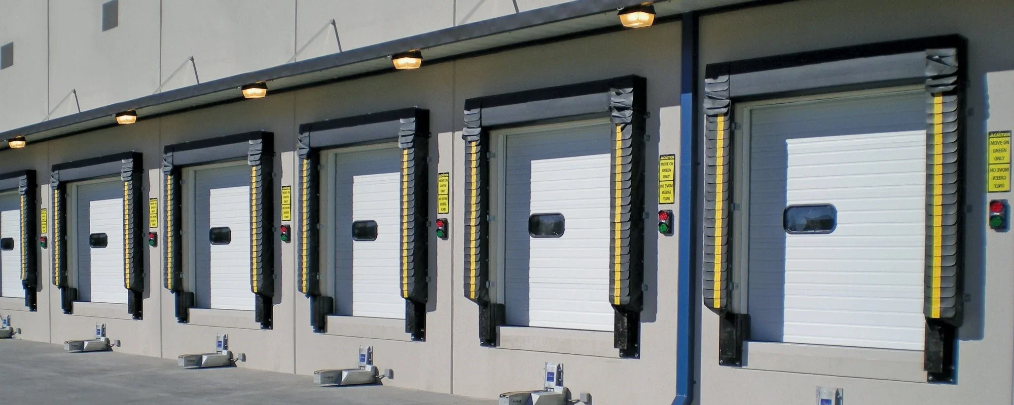 A row of multiple industrial loading dock doors with yellow and black safety barriers, red and green alert lights, and small windows in the center, attached to a warehouse or storage facility.
