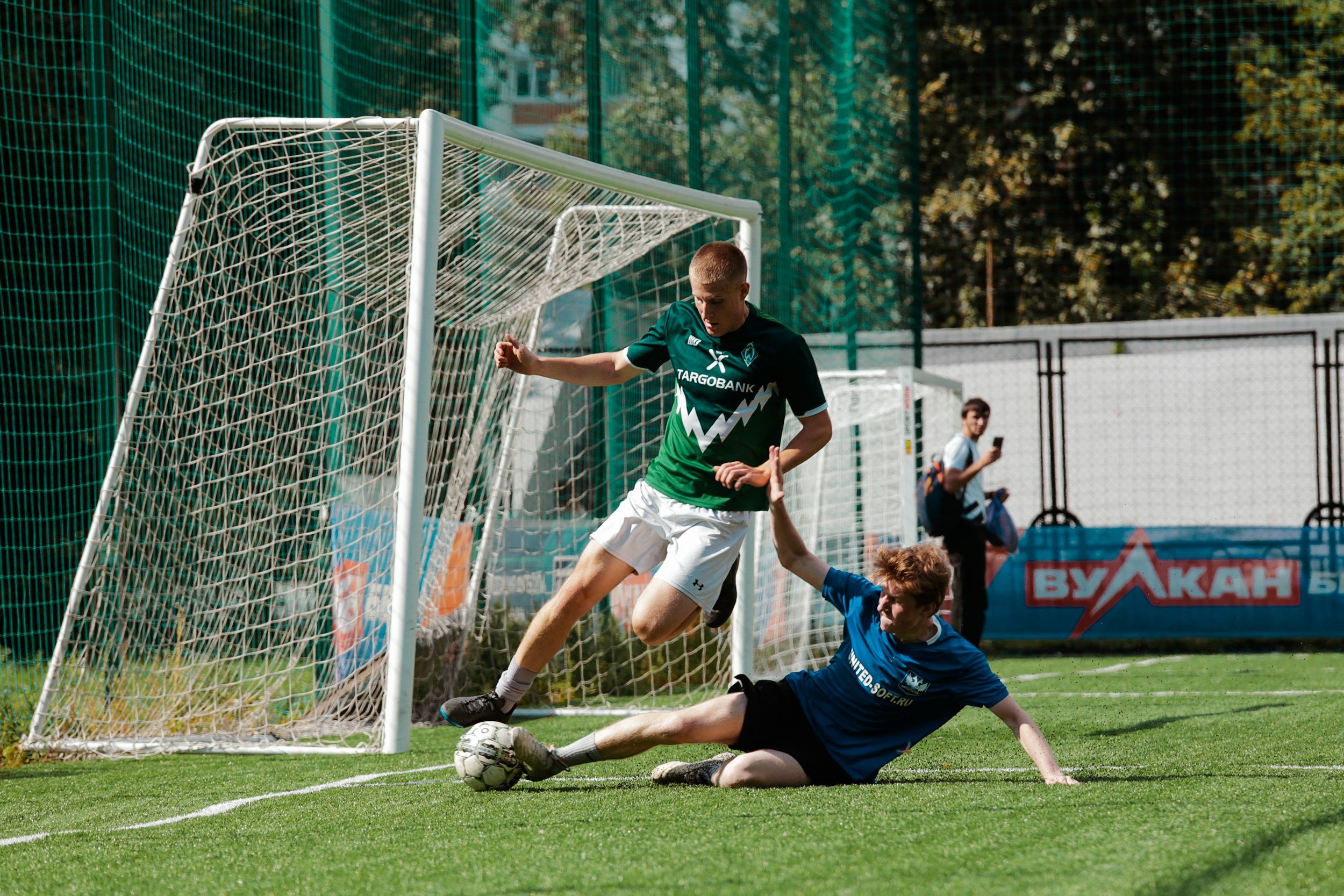 A soccer player in green jersey attempts to score a goal while a goalkeeper in blue dives to block the shot on a green field during daytime.