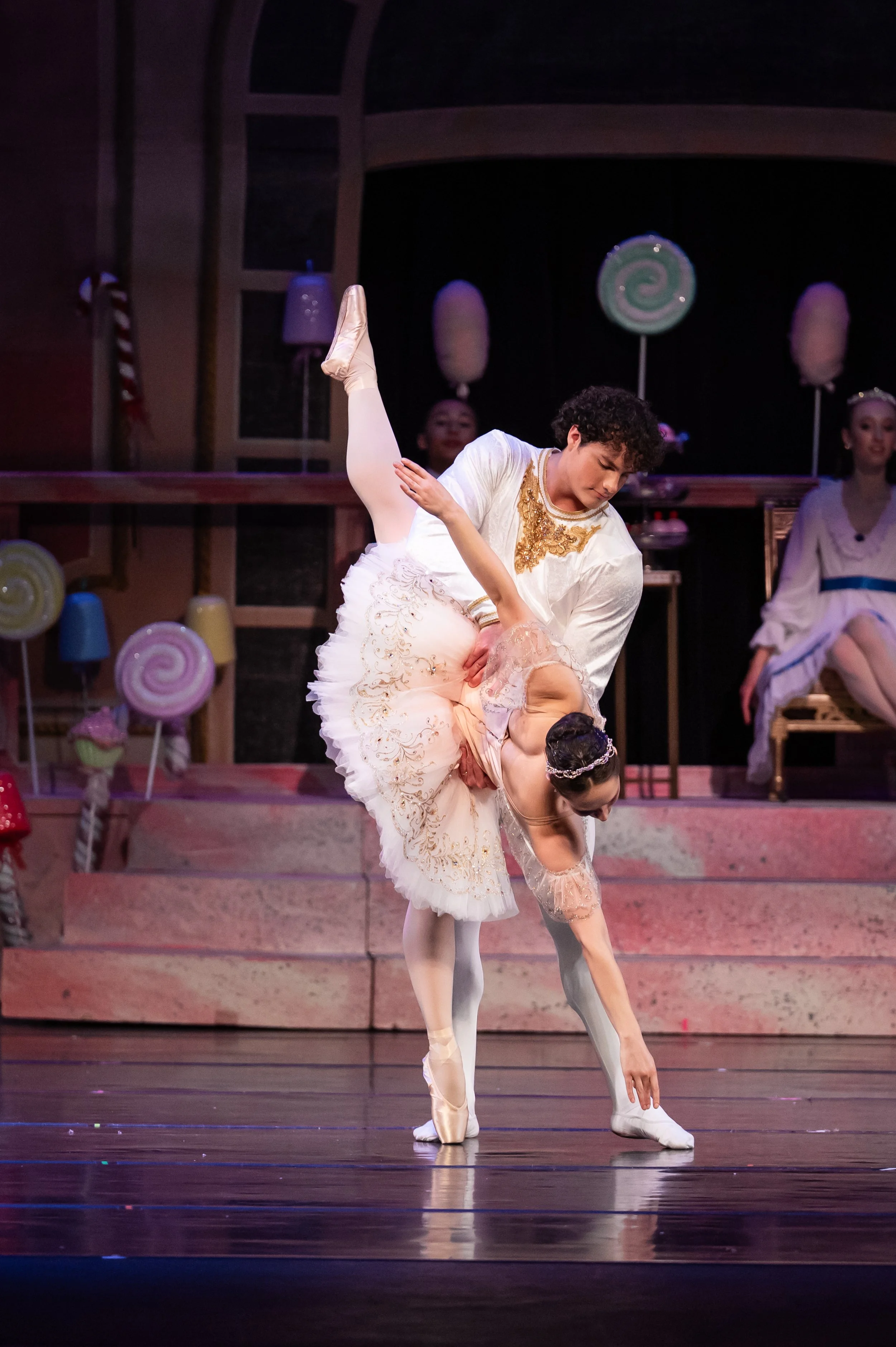 A male ballet dancer with curly hair performing a pose with a female ballet dancer in a tutu. The male dancer stands on one foot with the other leg lifted and bent, supporting the female dancer who is bent forward touching her toes. Both are on stage with colorful candy-themed props and other performers in costumes in the background.