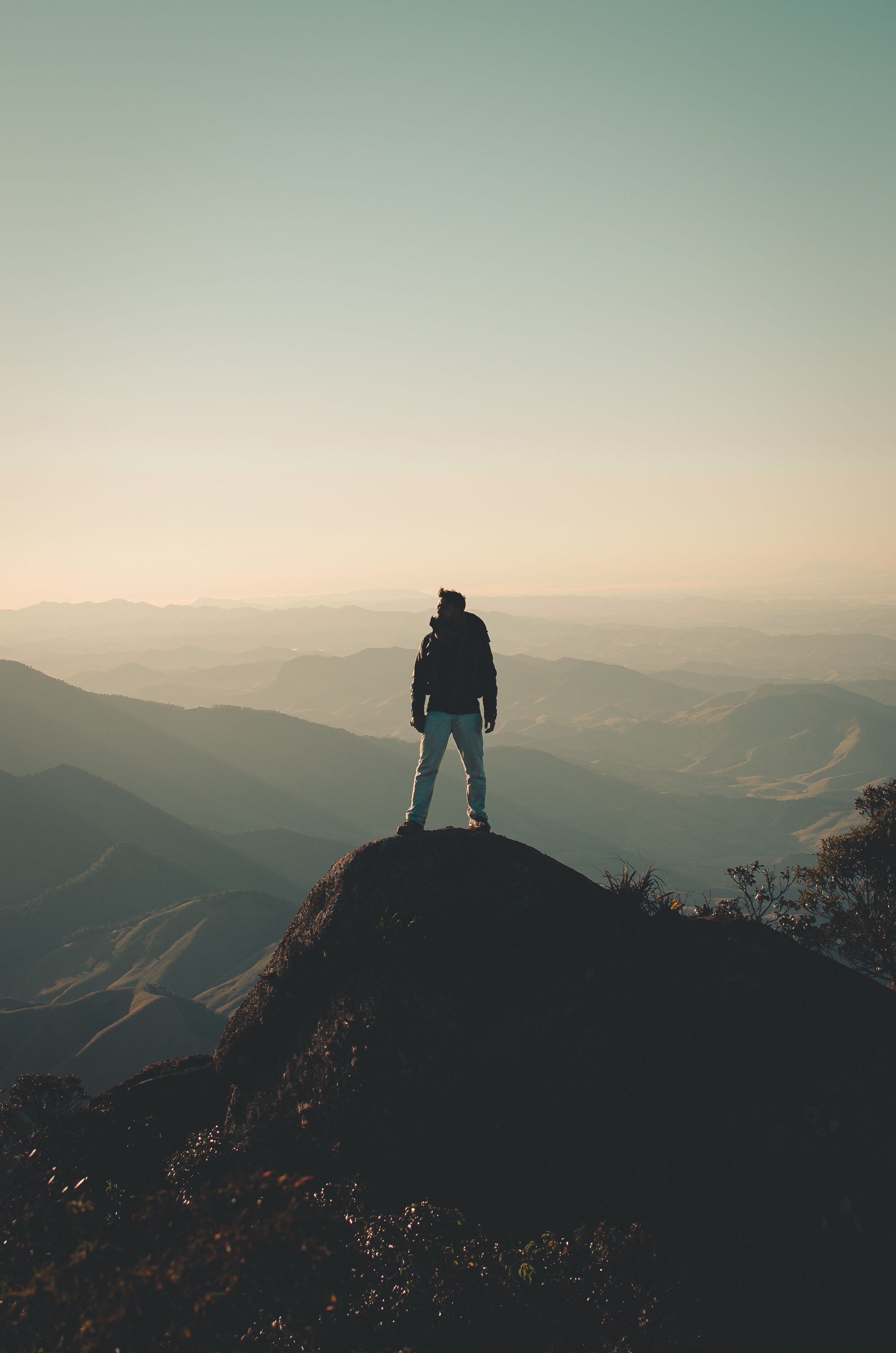 Silhouette of a person standing on a large rock at the top of a mountain during sunset, overlooking distant mountain ranges and valleys, with a clear sky.