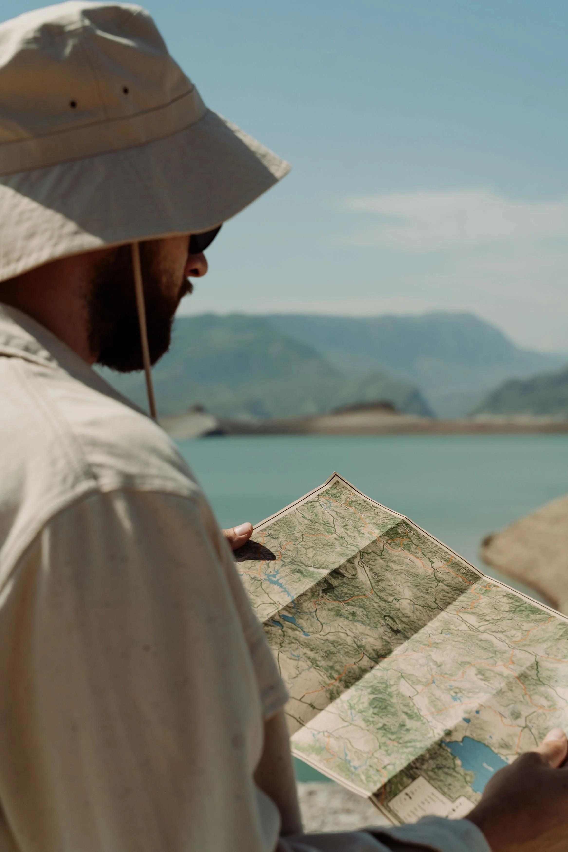 A person wearing a wide-brimmed hat and light clothing is holding and looking at a folded map outdoors, with a border of water and mountains in the background.