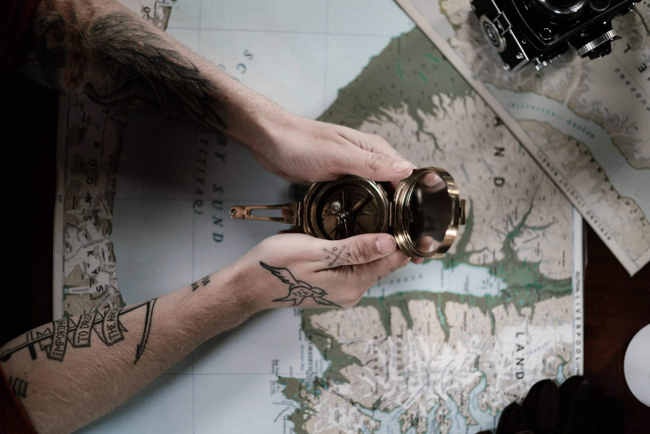 A person's arms with tattoos holding a brass compass over a detailed topographic map of a coastal area.