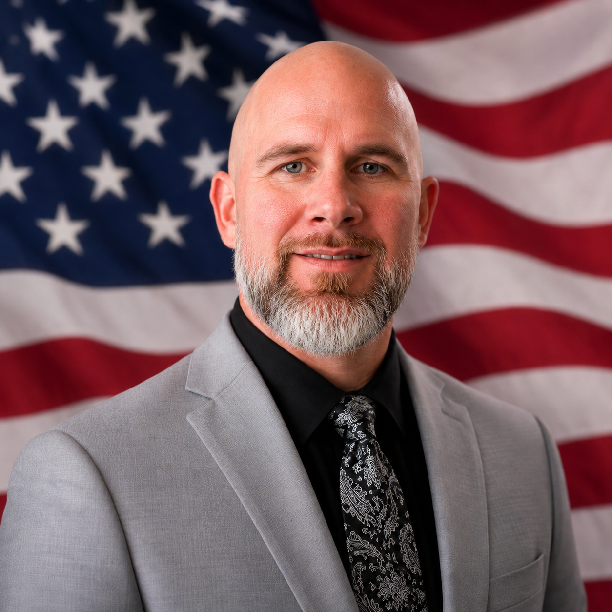 Portrait of a man with a gray suit, black shirt, and patterned tie, standing in front of an American flag.