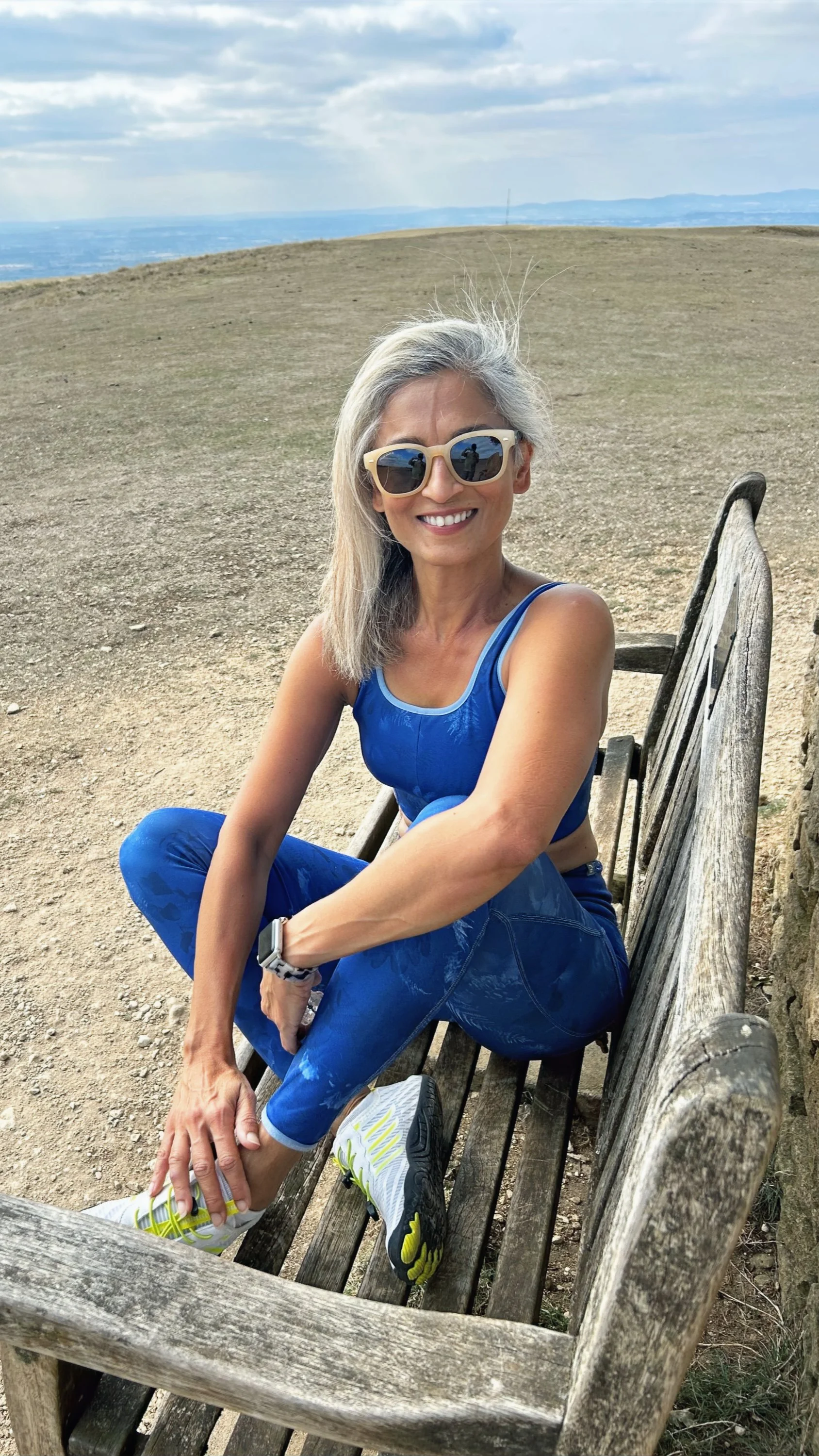 Smiling woman with gray hair, wearing sunglasses, a blue sports tank top and leggings, sitting on a wooden park bench on a hilltop with a cloudy sky and distant landscape in the background.