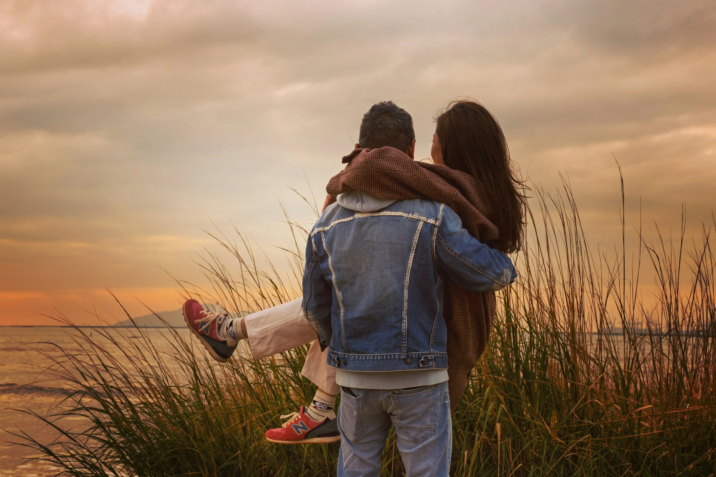 A couple standing in a grassy field near a body of water at sunset, with the woman giving the man a piggyback ride, both looking at each other.