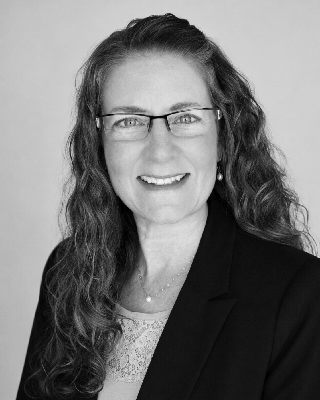 Black and white portrait of a smiling woman with curly hair, glasses, and a pearl earring, wearing a blazer and lace top, against a plain background.