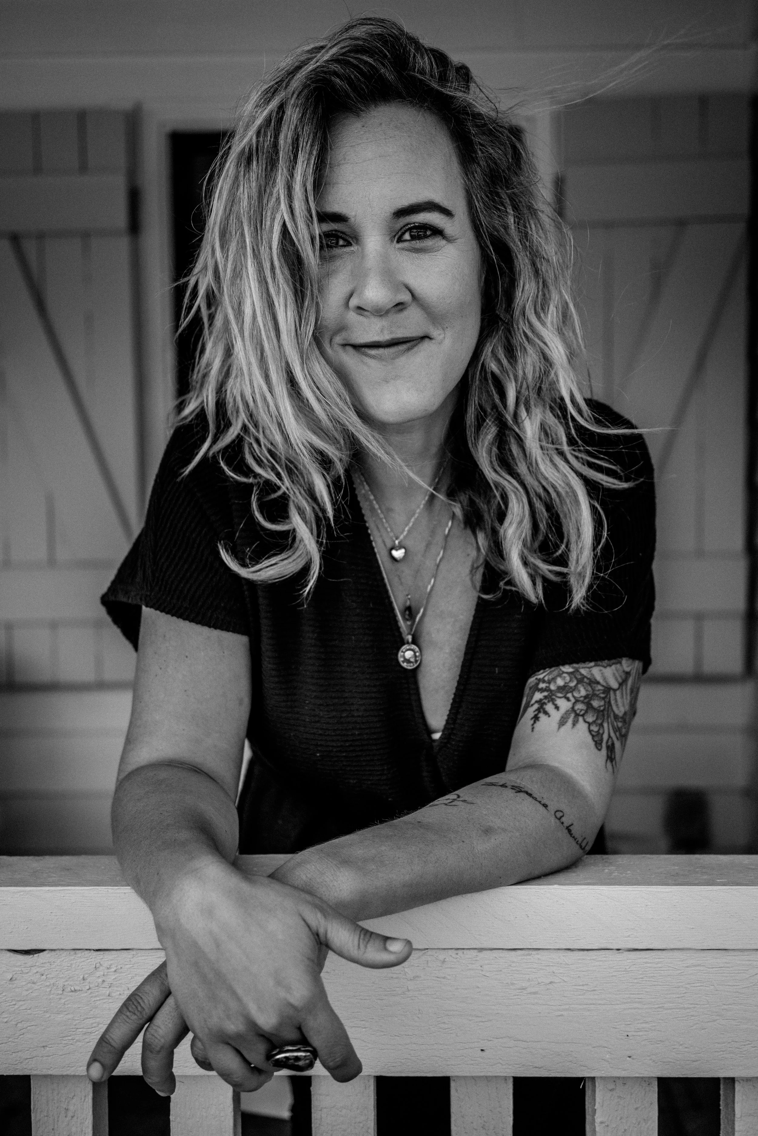 Black and white portrait of a smiling woman with wavy hair, wearing necklaces and a short-sleeved shirt, sitting at a wooden table with her hands folded.