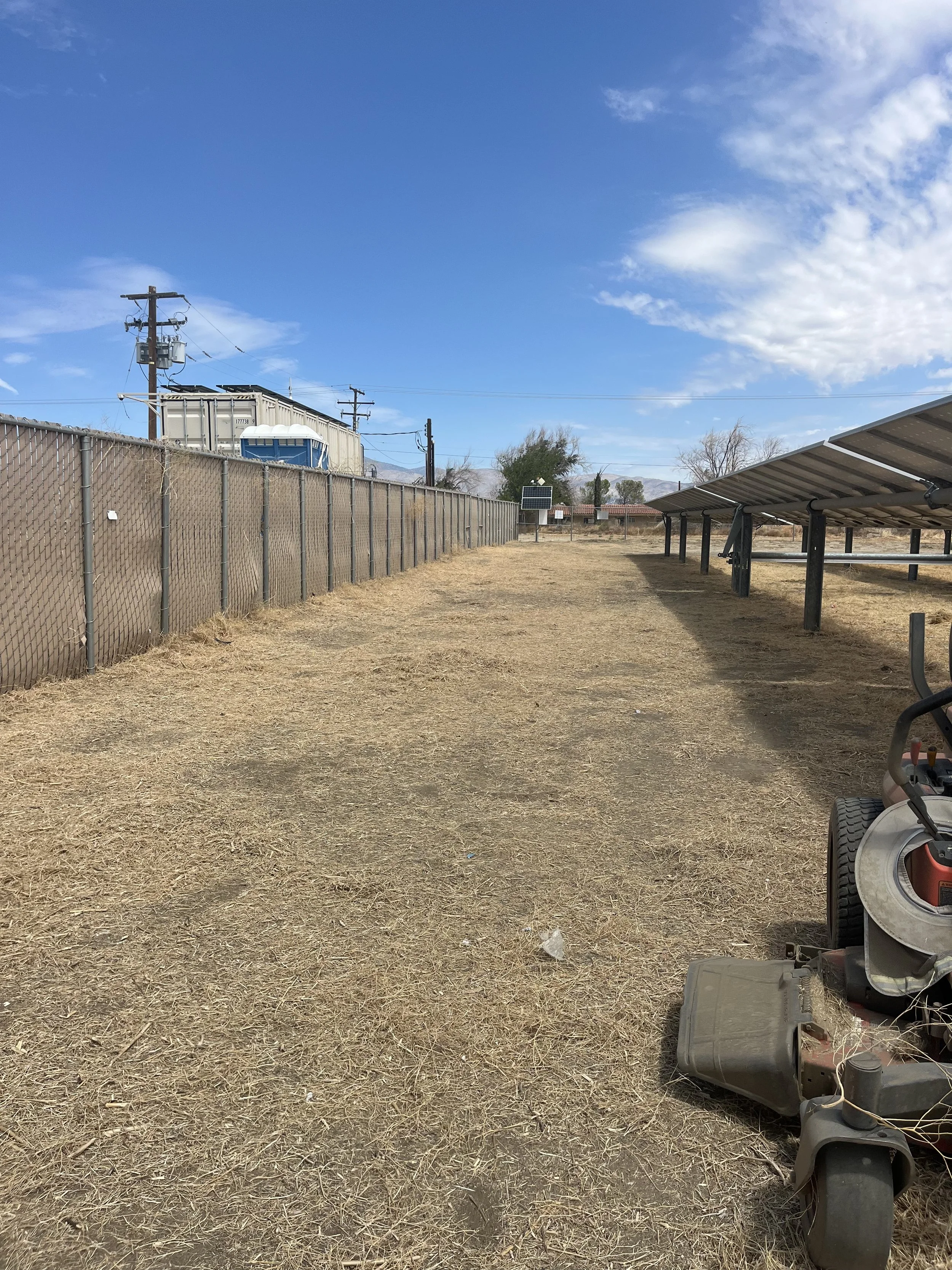 Open outdoor area with solar panels on the right, a chain-link fence on the left, portable toilets, utility pole with wires, dry grass ground, and a blue sky with some clouds.