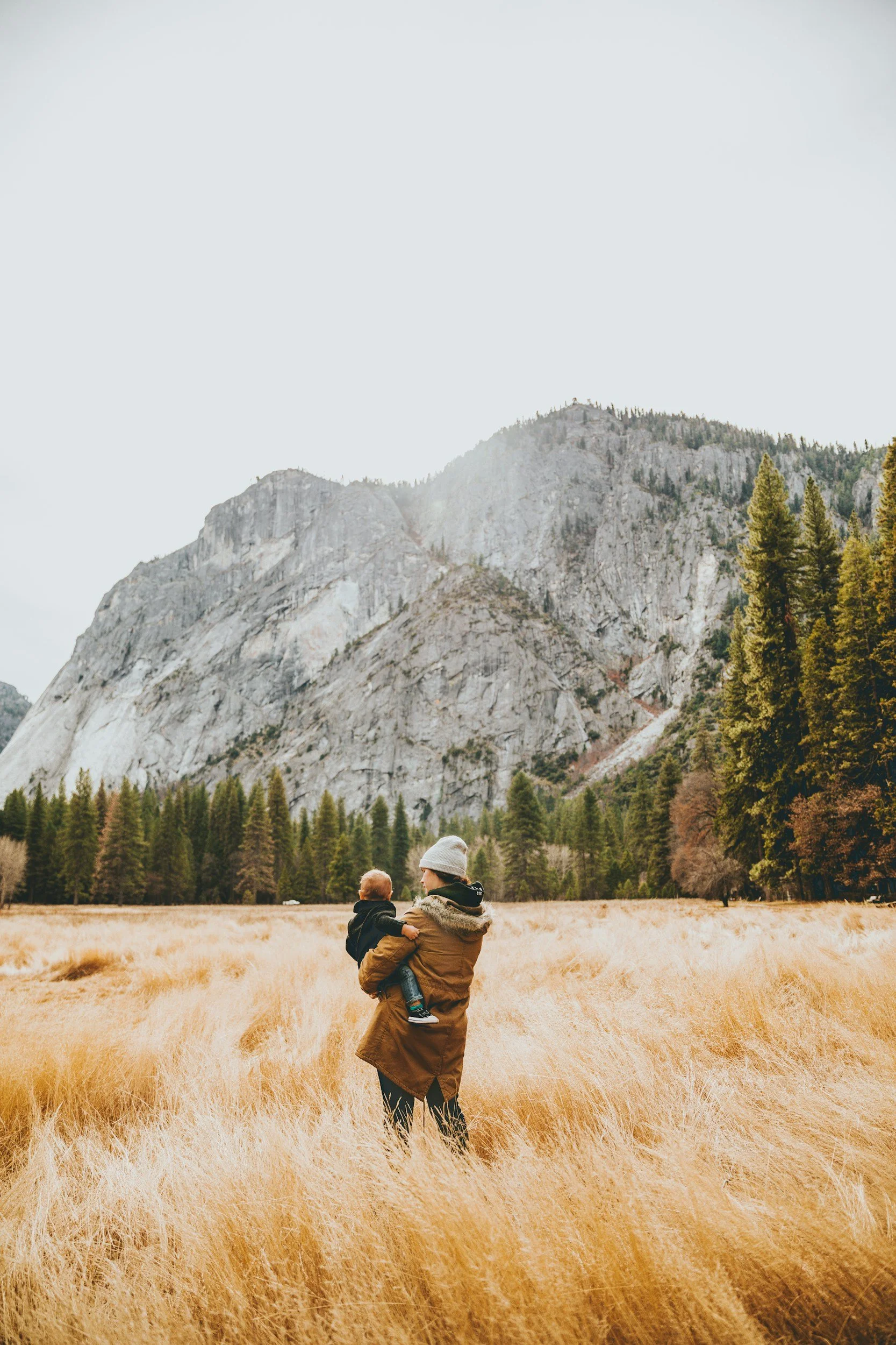 A person carrying a child in a yellow field with tall grass, mountains, and trees in the background during daylight.
