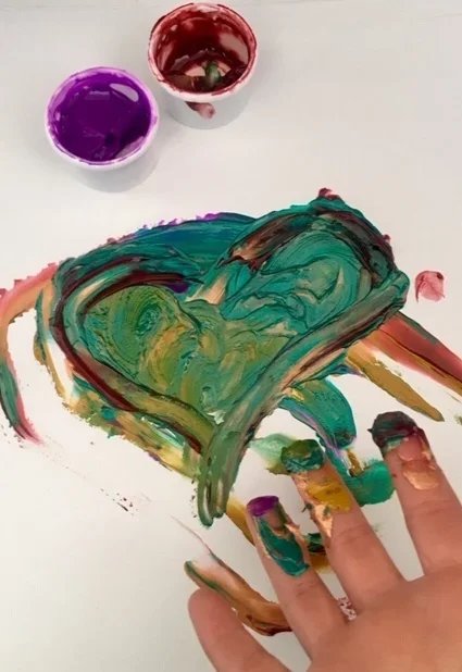 Child's hand covered in multicolored paint creating artwork on paper with two cups of purple and red paint nearby.