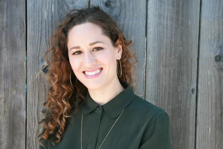 Smiling woman with curly hair, wearing a dark green shirt, hoop earrings, and a gold necklace, standing in front of a wooden fence.