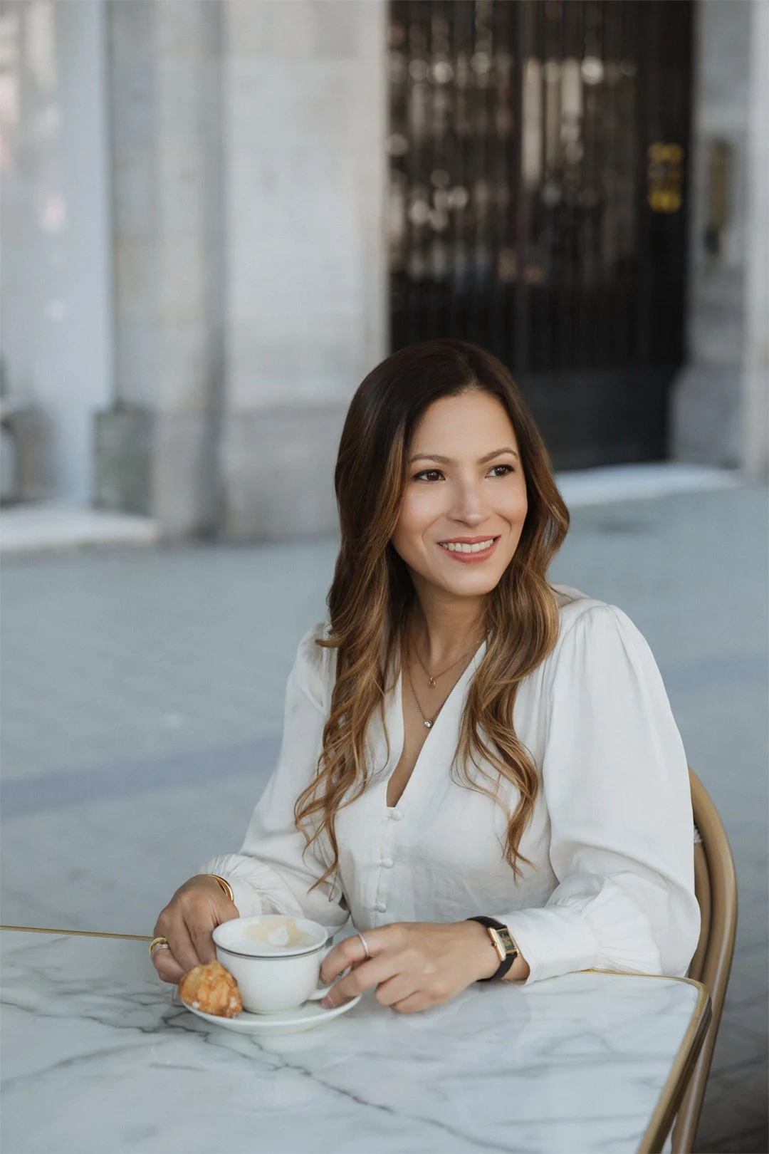 A woman with wavy brown hair sitting at a marble table, holding a cup of coffee or tea, smiling, in an indoor setting with a modern, industrial background. Bianca Inoa Business Transformation & Strategic Advisor.