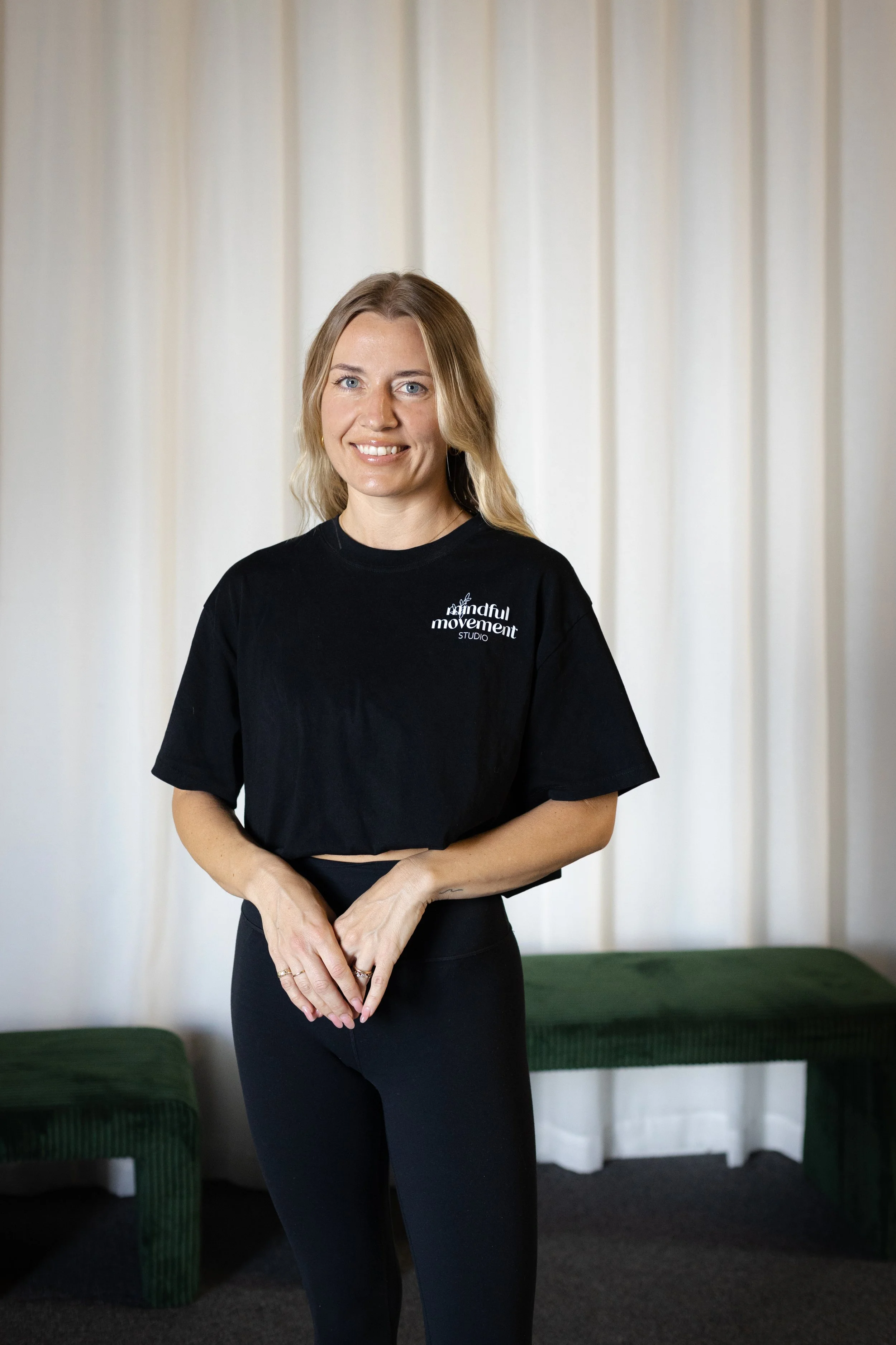 A smiling woman with blonde hair, wearing a black T-shirt with the logo 'mindful movement studio,' stands in front of white curtains with green furniture behind her.