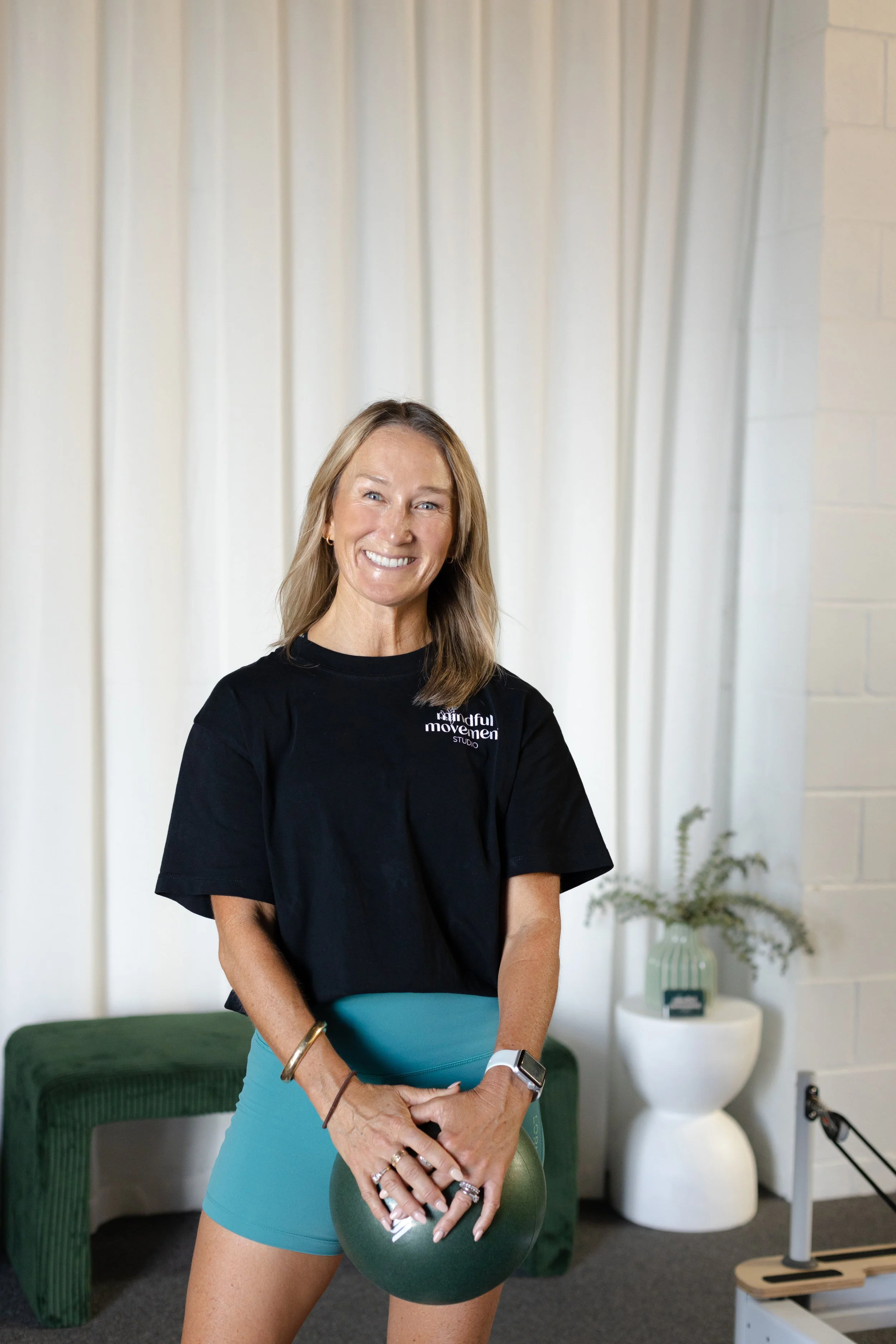 A smiling woman in workout attire holding a green kettlebell in a gym or fitness studio. She has long blonde hair, wears a black t-shirt with white text, blue workout shorts, and several rings and bracelets. There is a green bench, a white table with a vase of flowers, and various fitness equipment in the background.