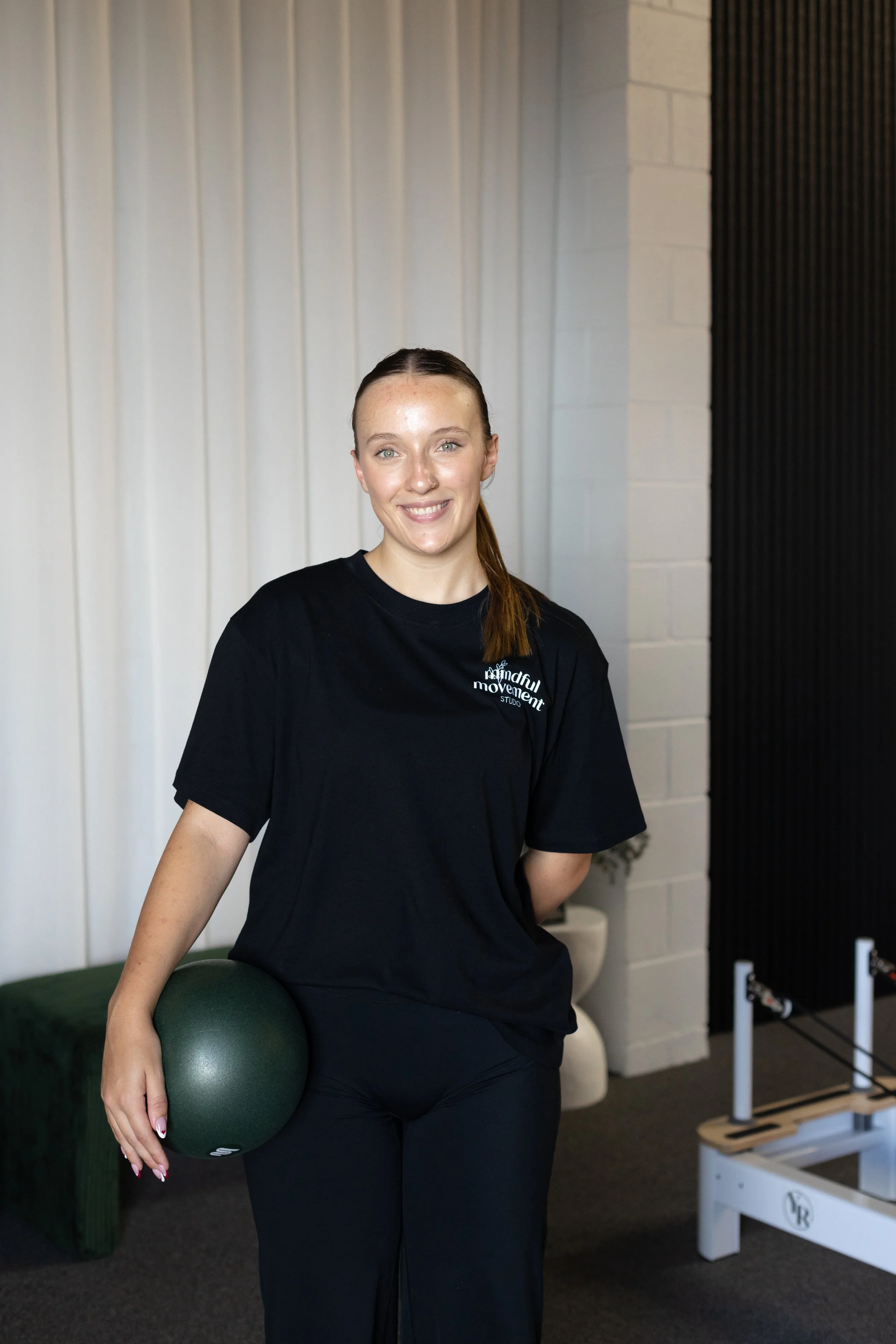 A woman smiling at the camera, holding a green exercise ball against her right hip, in a room with white and black walls, a green bench, and some fitness equipment.