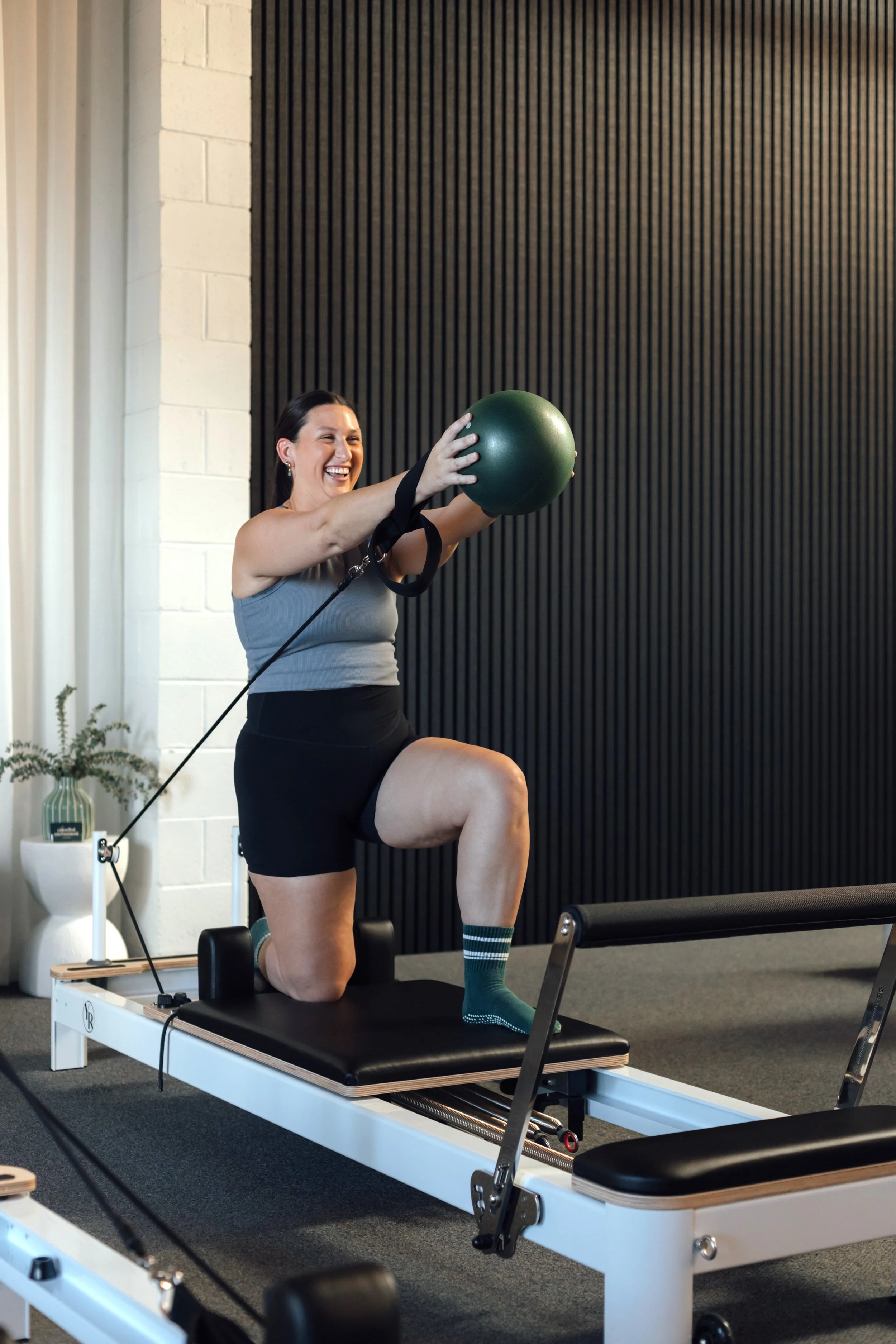Mindful Movement Studio client is on a Pilates Reformer, holding a green Pilates ball with a joyful expression.