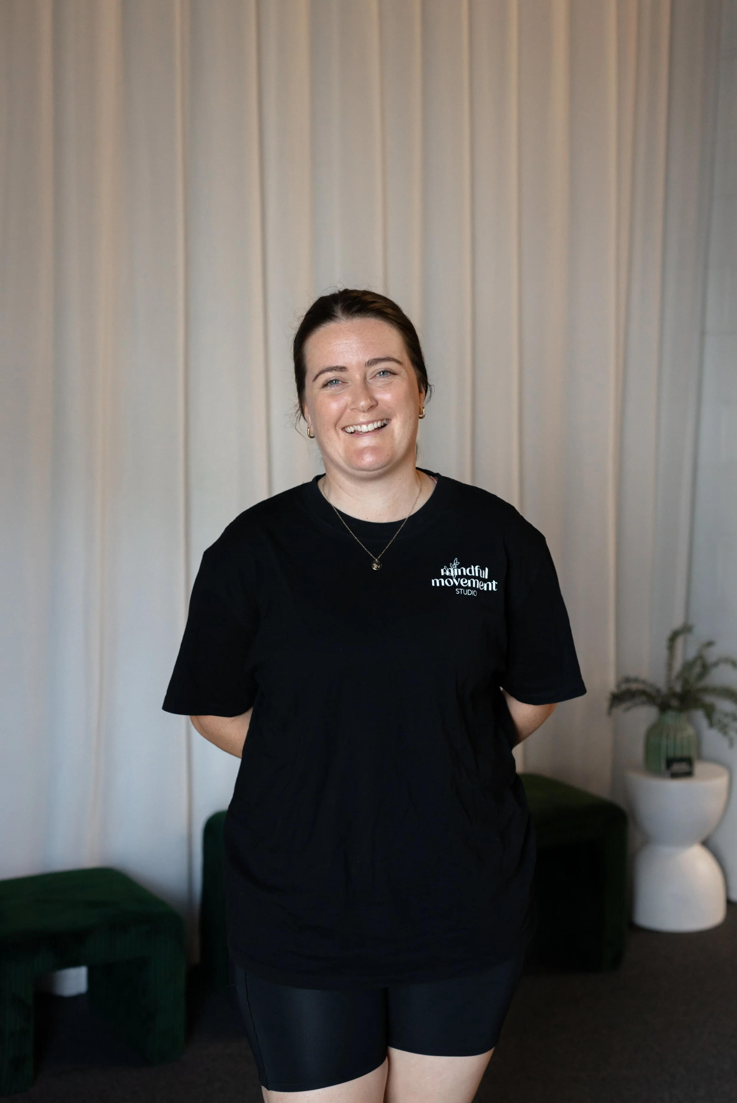 A woman with dark hair, wearing earrings, a necklace, and a black T-shirt, standing in front of a beige curtain, smiling at the camera. The T-shirt has white text that reads 'mindful movement studio'.