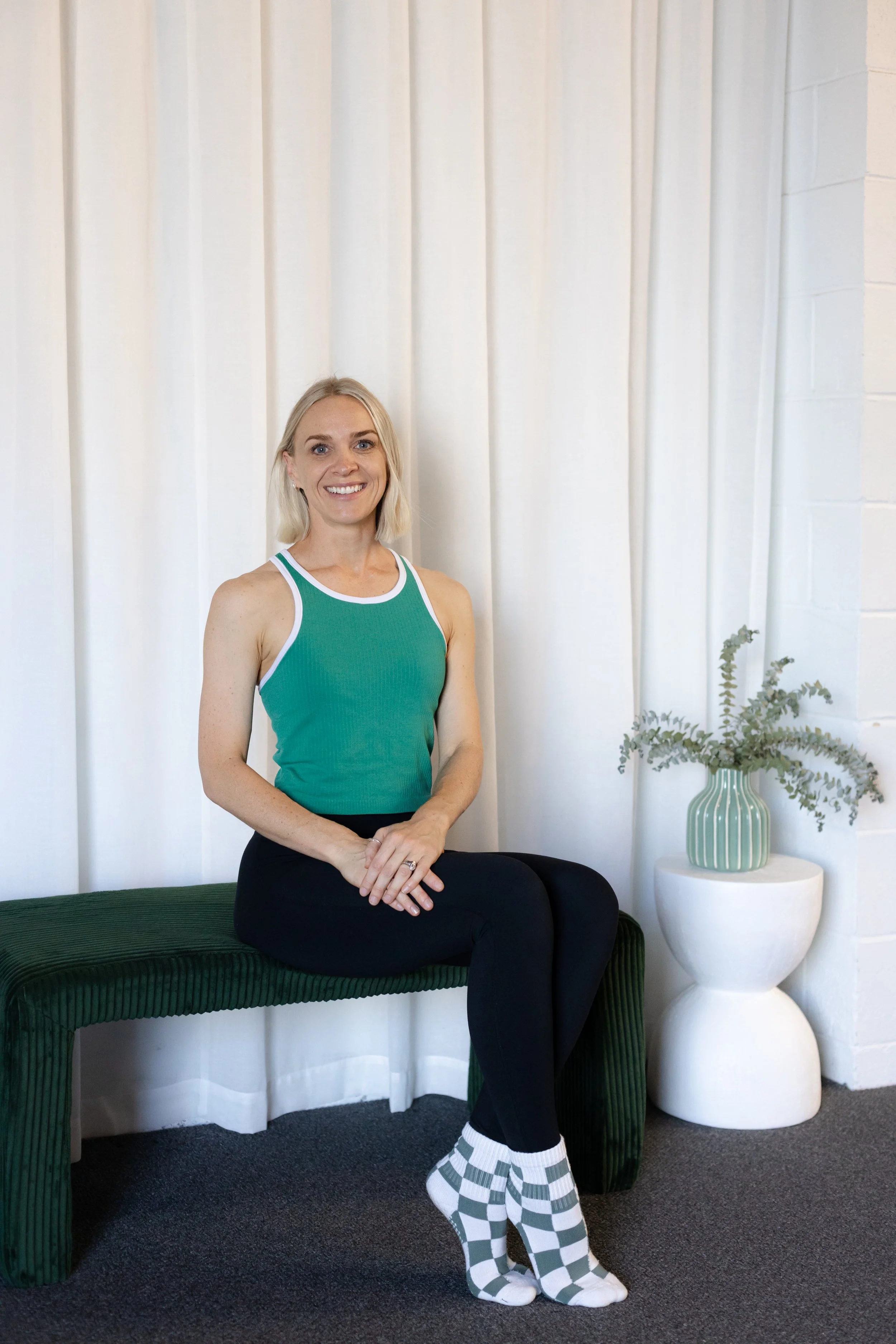 A woman with blond hair and a green tank top, sitting on a green velvet bench. Behind her is a white curtain and next to her is a side table with a striped green and white vase filled with foliage.