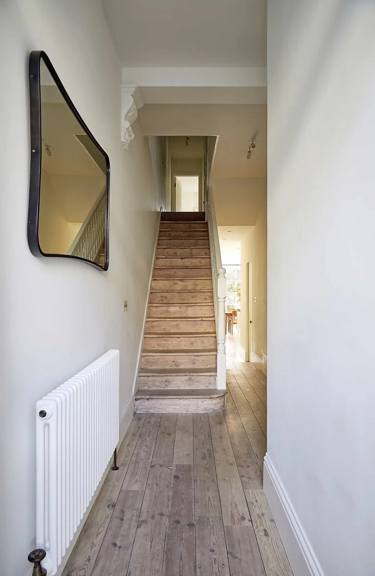 Interior view of a hallway with wooden floors, a staircase leading upstairs, a white radiator on the left wall, a rectangular mirror on the left, and partial views of rooms and a dining area in the background.