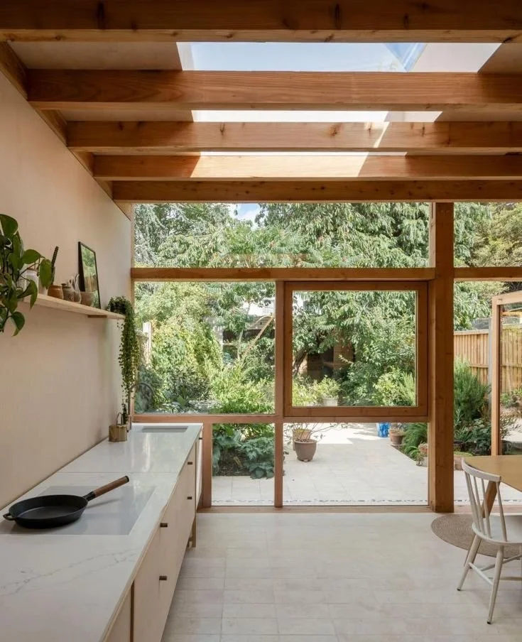 View of a modern kitchen with wooden beams, white cabinetry, and large windows overlooking a lush garden.