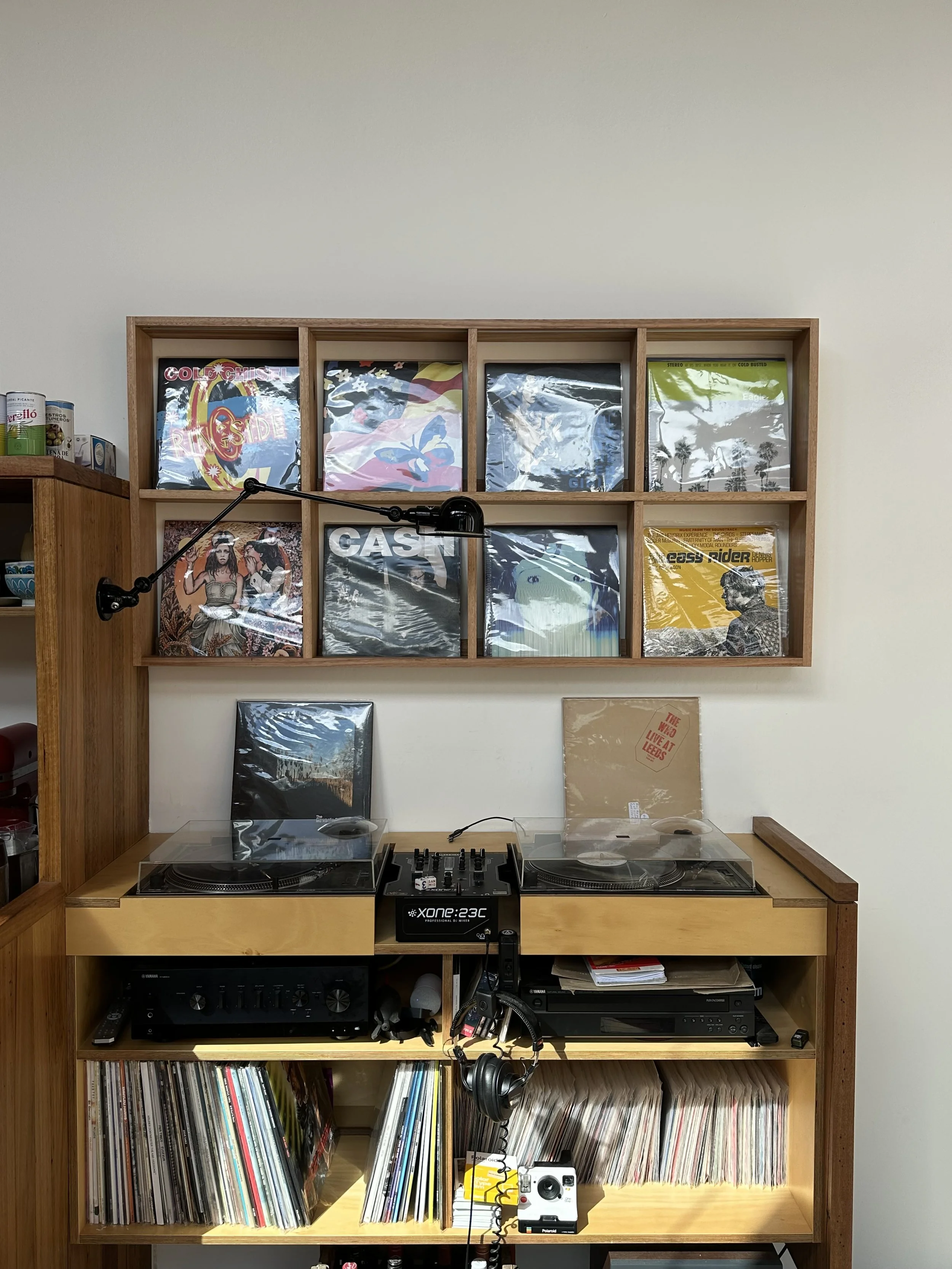 A wooden shelf with a collection of vinyl records in cover art sleeves, a DJ turntable setup with headphones hanging, and a stereo receiver below. The background features a plain wall and a smaller shelf with canned goods and decorative items.