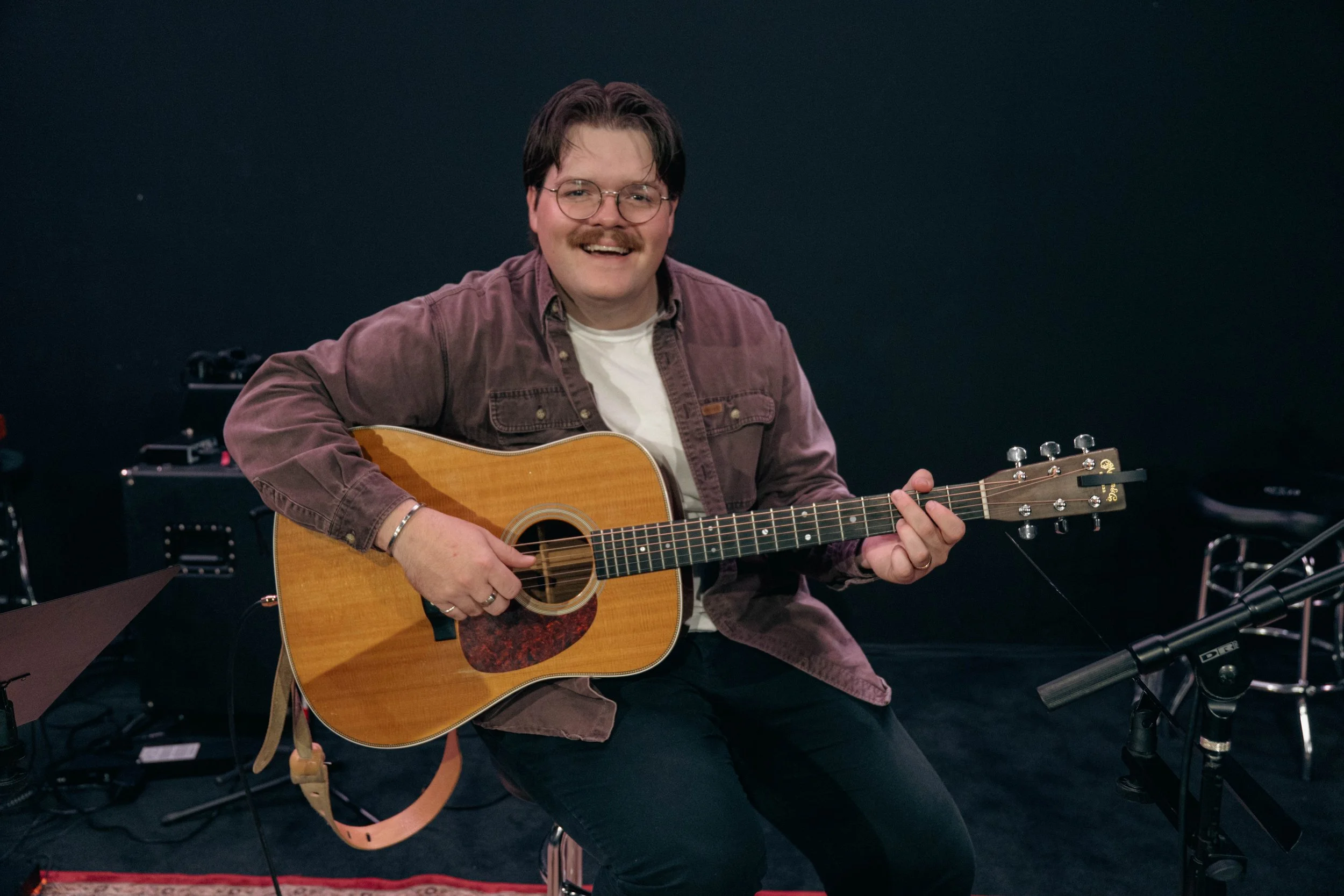 A music teacher playing an acoustic guitar, sitting in a music studio with equipment around him.