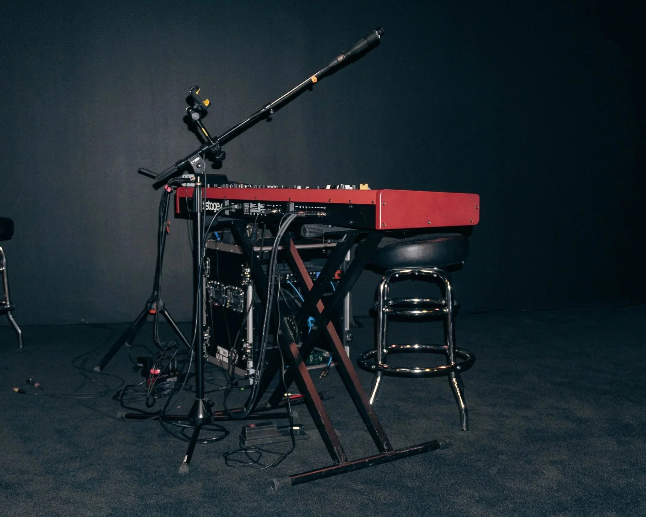 A musical keyboard with electronic and audio equipment on a stand, along with a drum stool, set against a dark background.