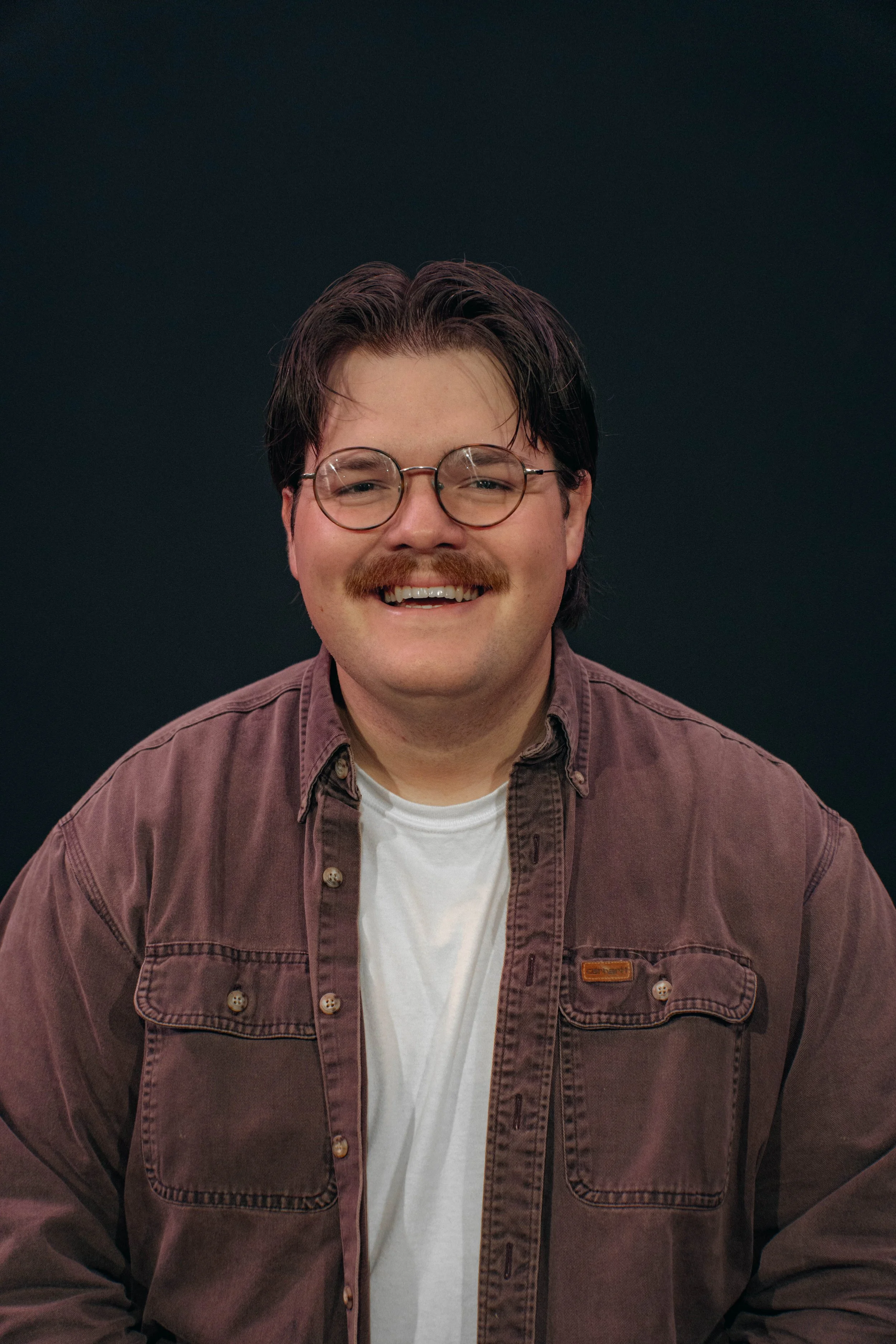A smiling man wearing glasses and a brown jacket over a white shirt, dark background