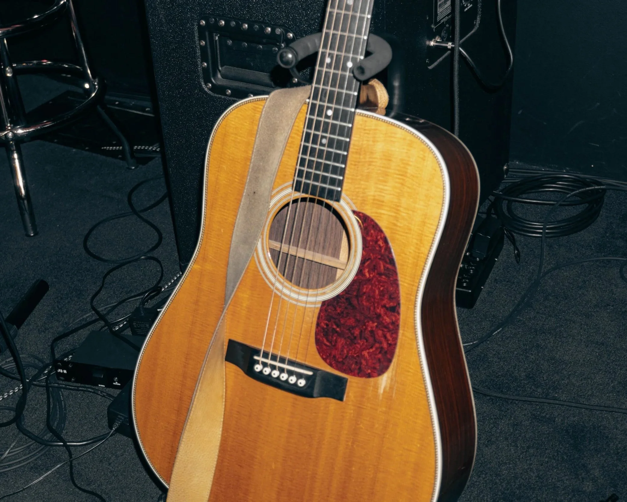 An acoustic guitar with a natural wood finish and a tortoiseshell pickguard leaning against a black amplifier, with various cords on the floor and a chrome stool nearby.