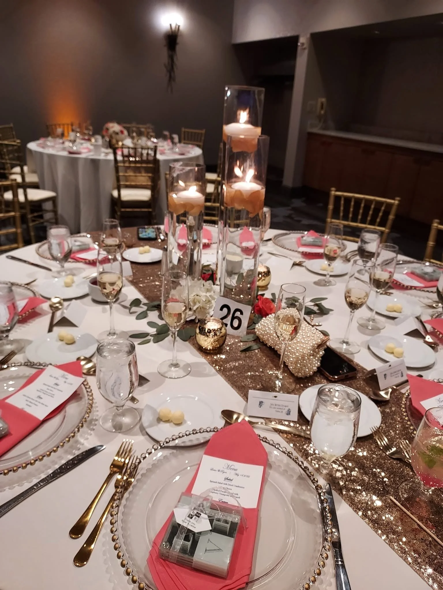 Elegant wedding reception table with gold and coral accents, tall glass candle holders, gold cutlery, and a menu card on each place setting.