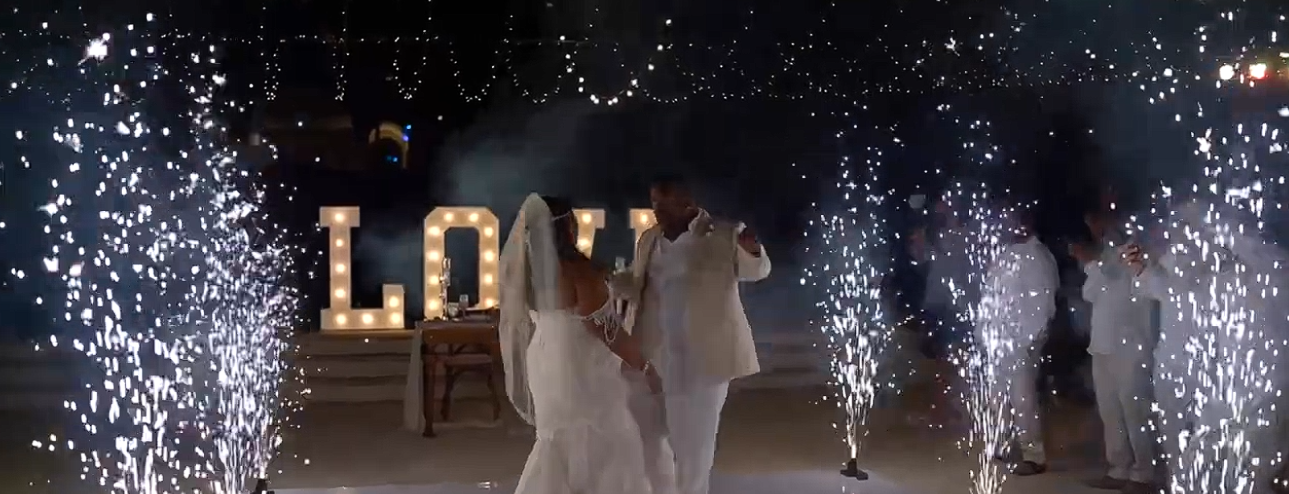 Bride and groom dancing beside indoor sparkler fountains and an illuminated LOVE sign at their wedding reception.