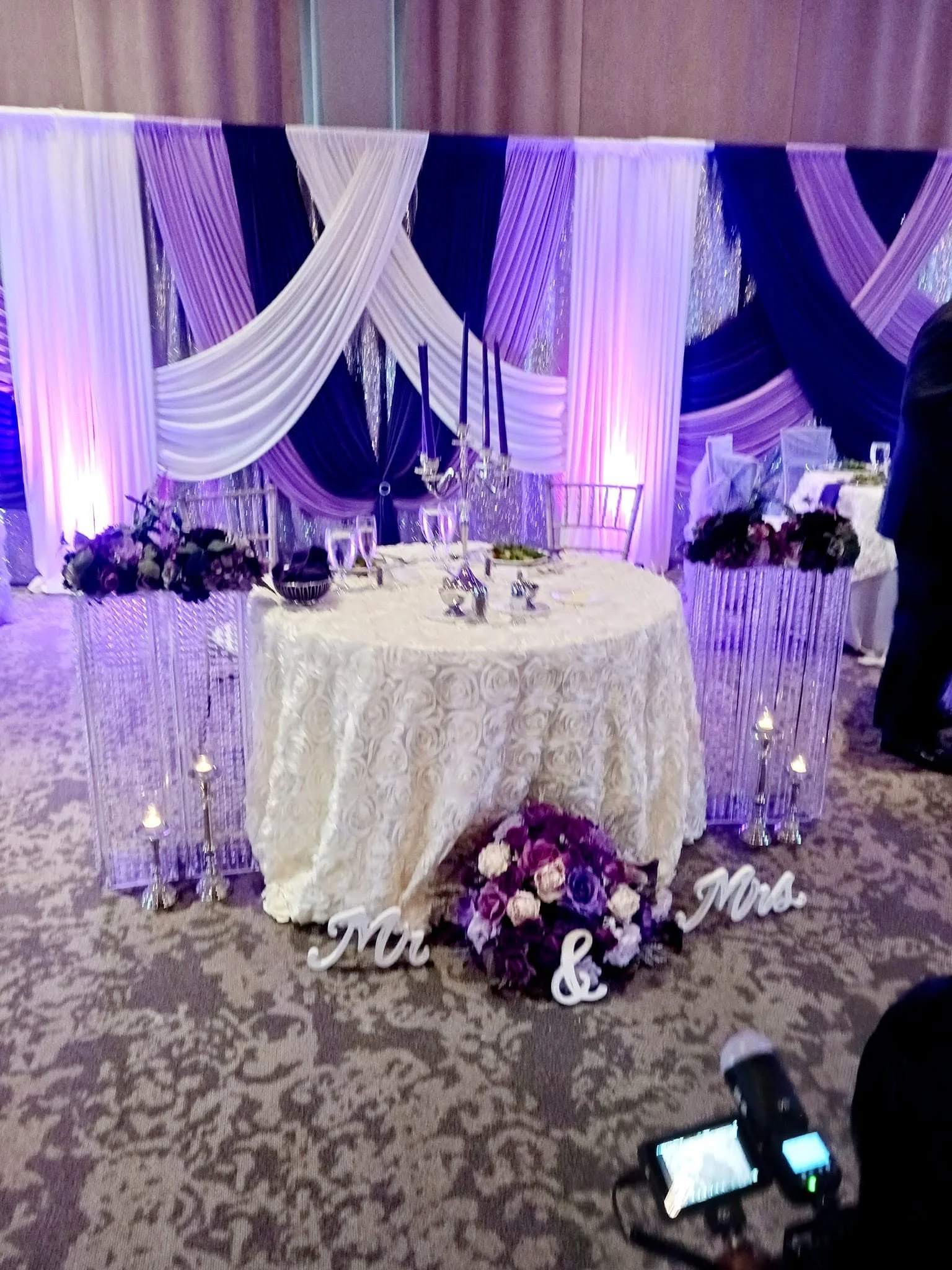 Decorative wedding or event table with white and purple drapery backdrop, floral arrangements, and sign reading 'Mr. & Mrs.'.