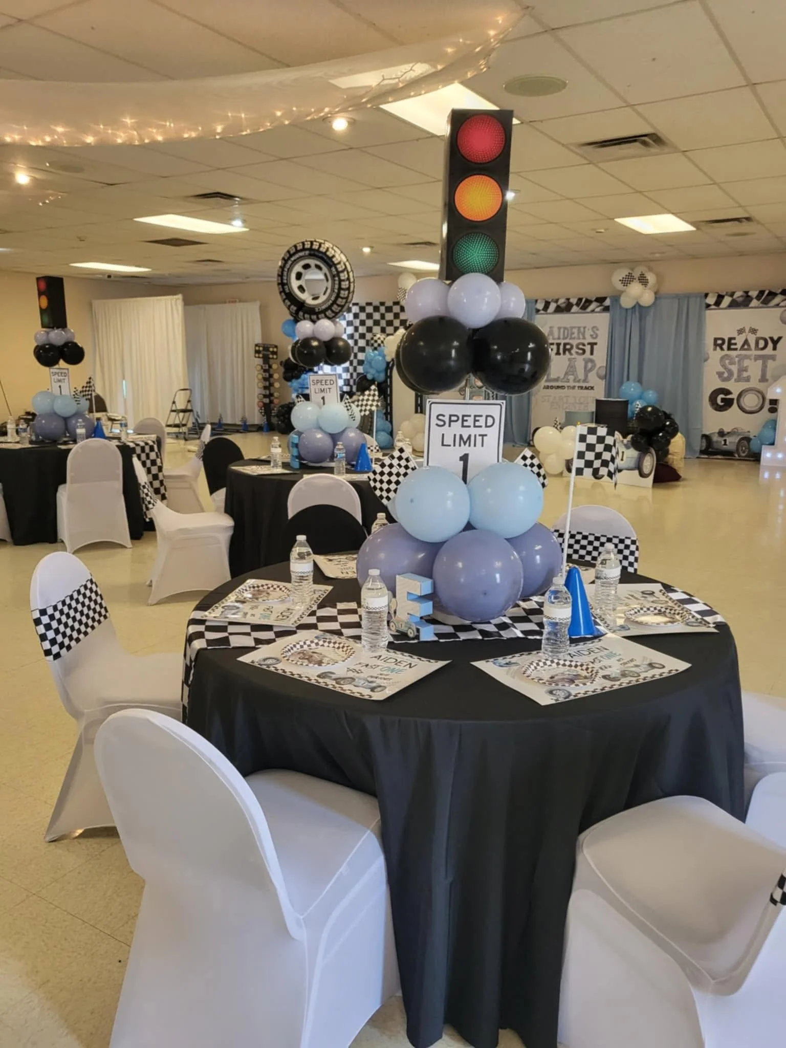 Race-car-themed party table with a checkered runner, black-and-blue balloons, a traffic-light centerpiece, custom water bottles, and party favors.