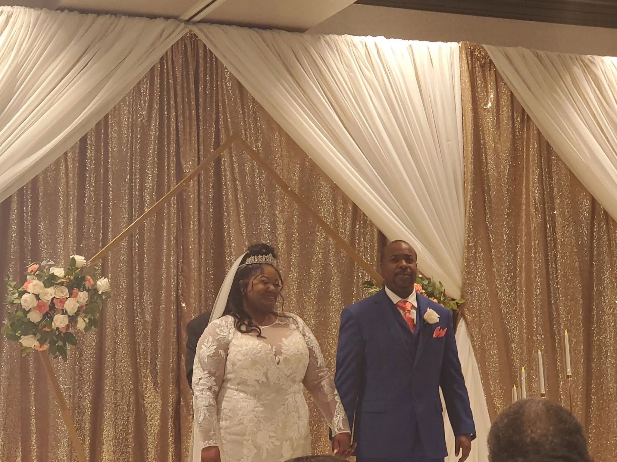 Bride and groom standing before a gold-and-cream draped ceremony backdrop with floral arrangements.