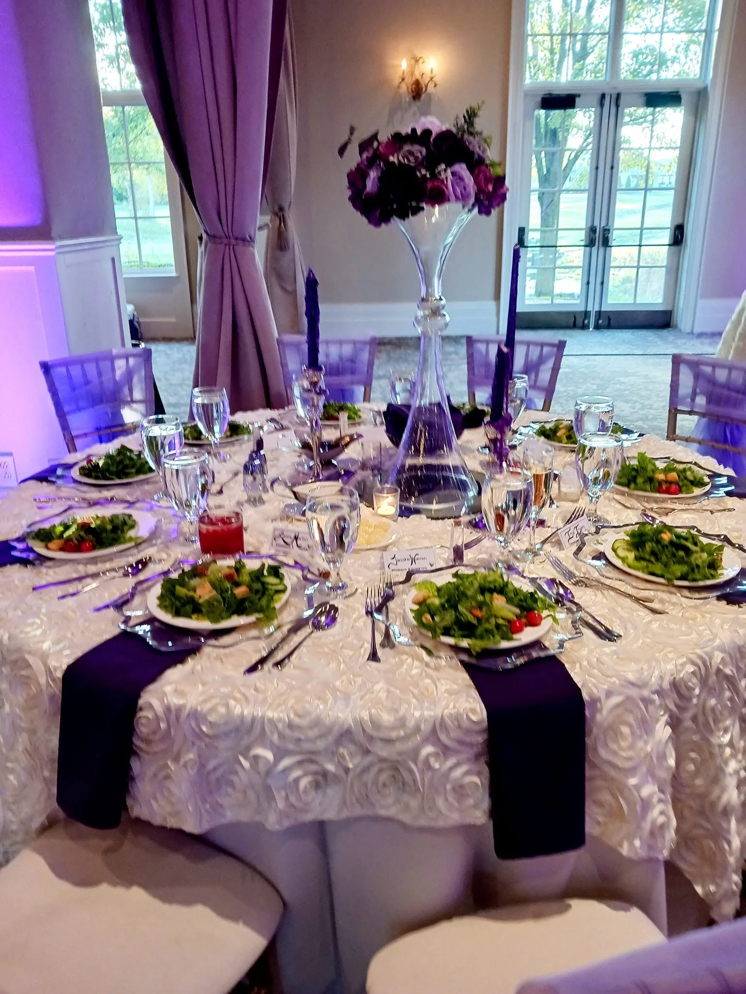 Formal banquet table with white textured linen, purple chair sashes, candles, place cards, and a purple-and-lavender floral centerpiece.