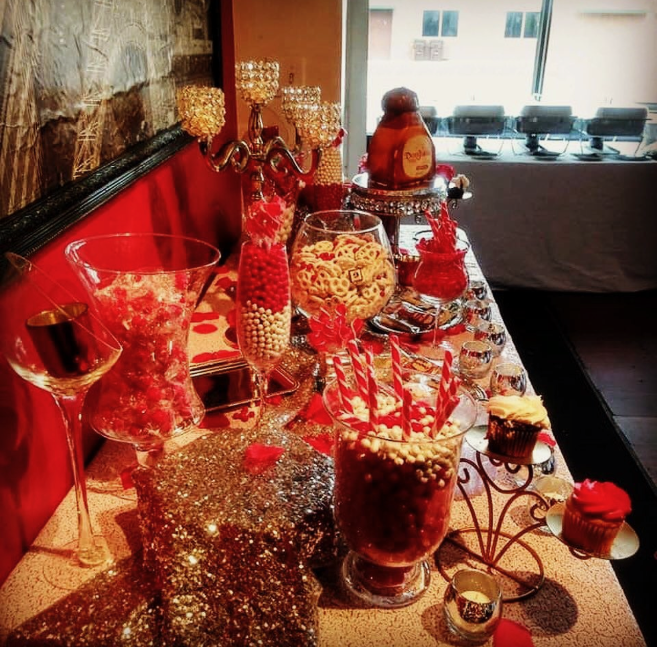 A table decorated for a celebration with red and gold accents, featuring various jars of candies, a chocolate fountain, a cupcake, and a large jar of Nutella. The table has a glittery gold cloth, and the background shows a room with a stone wall and 