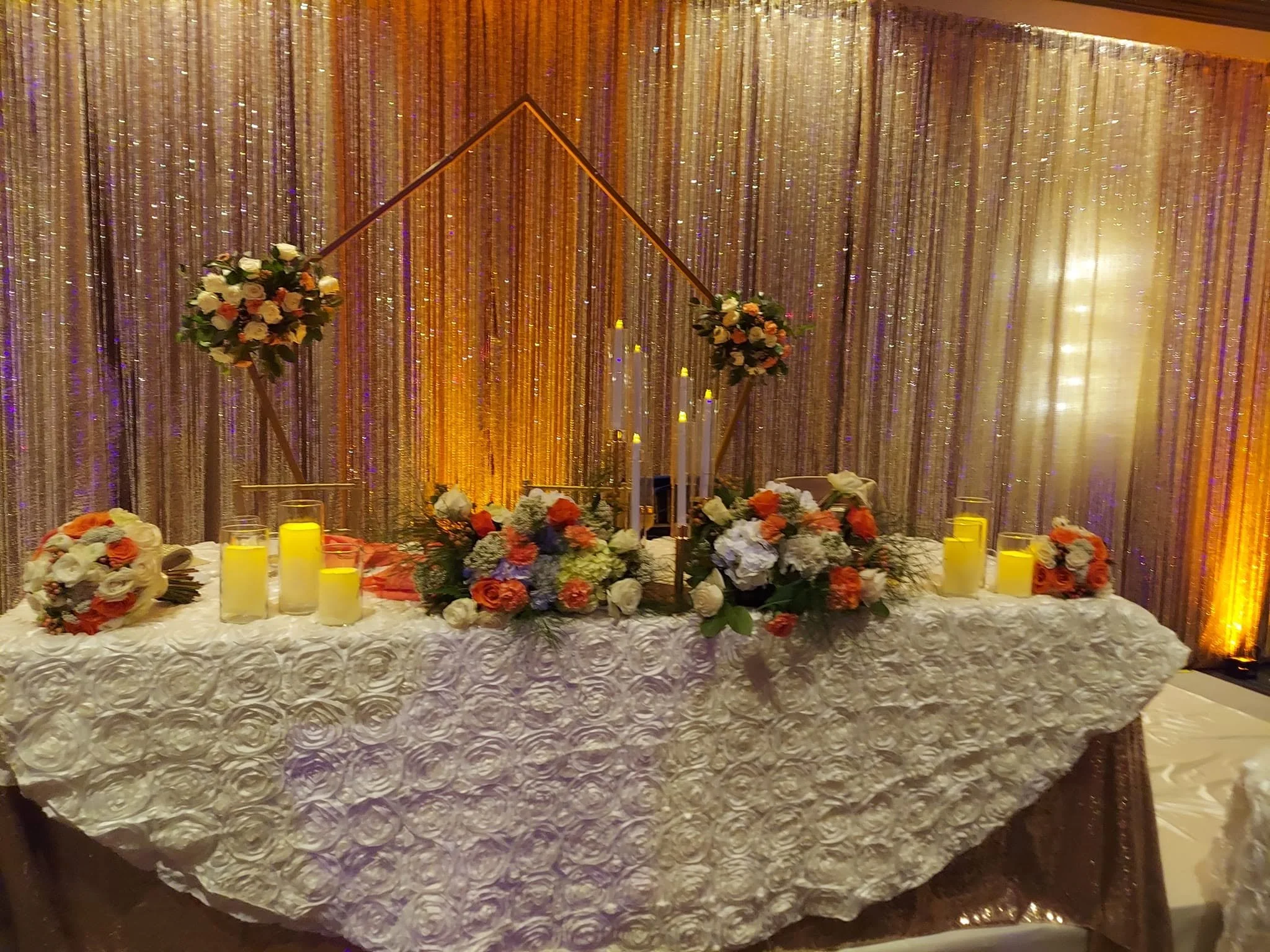 Wedding head table with white linens, floral arrangements, amber candles, and a shimmering gold and white backdrop.