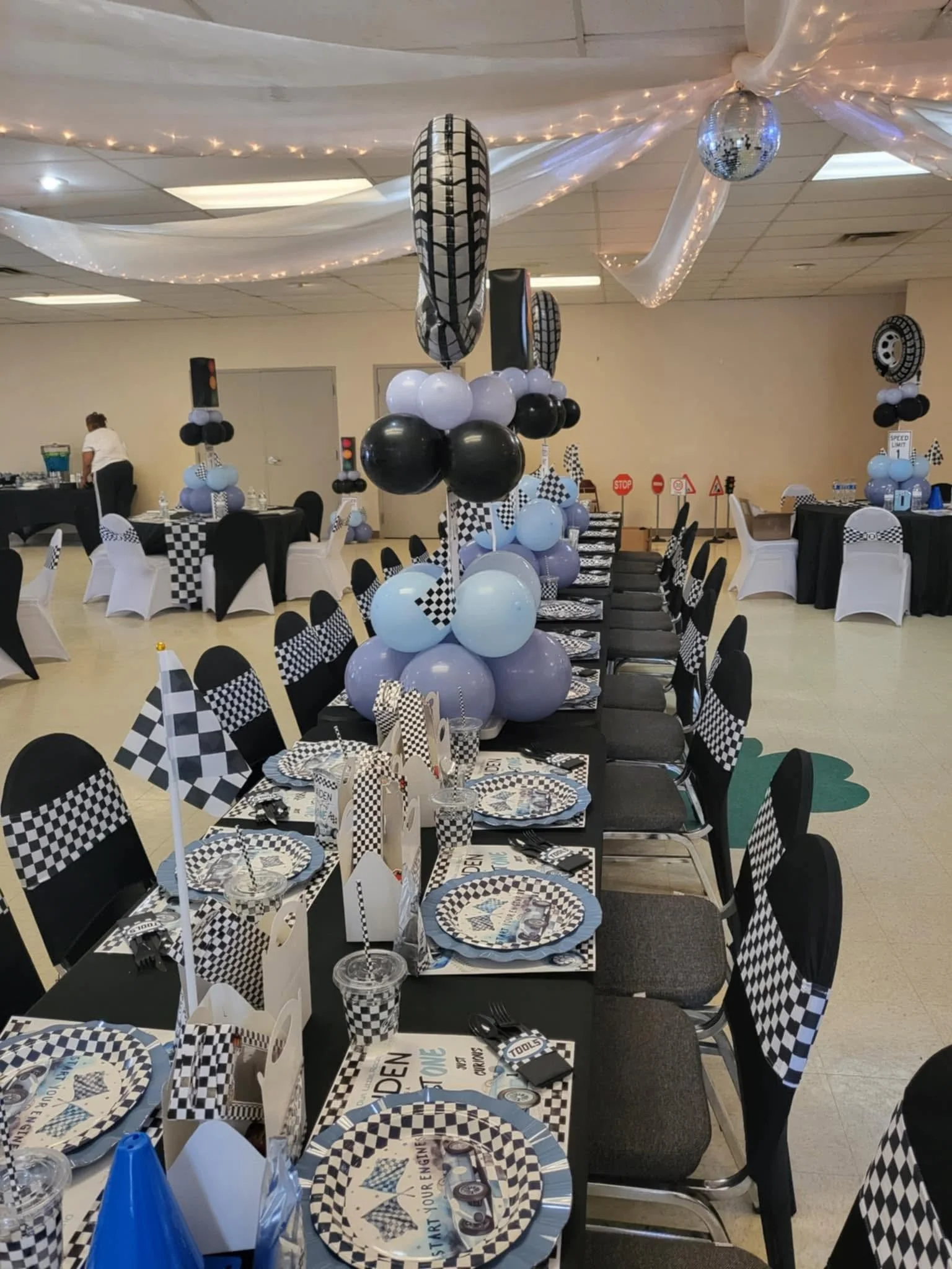Race-car-themed party room with black, white, and blue balloons, tire-pattern decorations, coordinated table settings, and checkered flags.