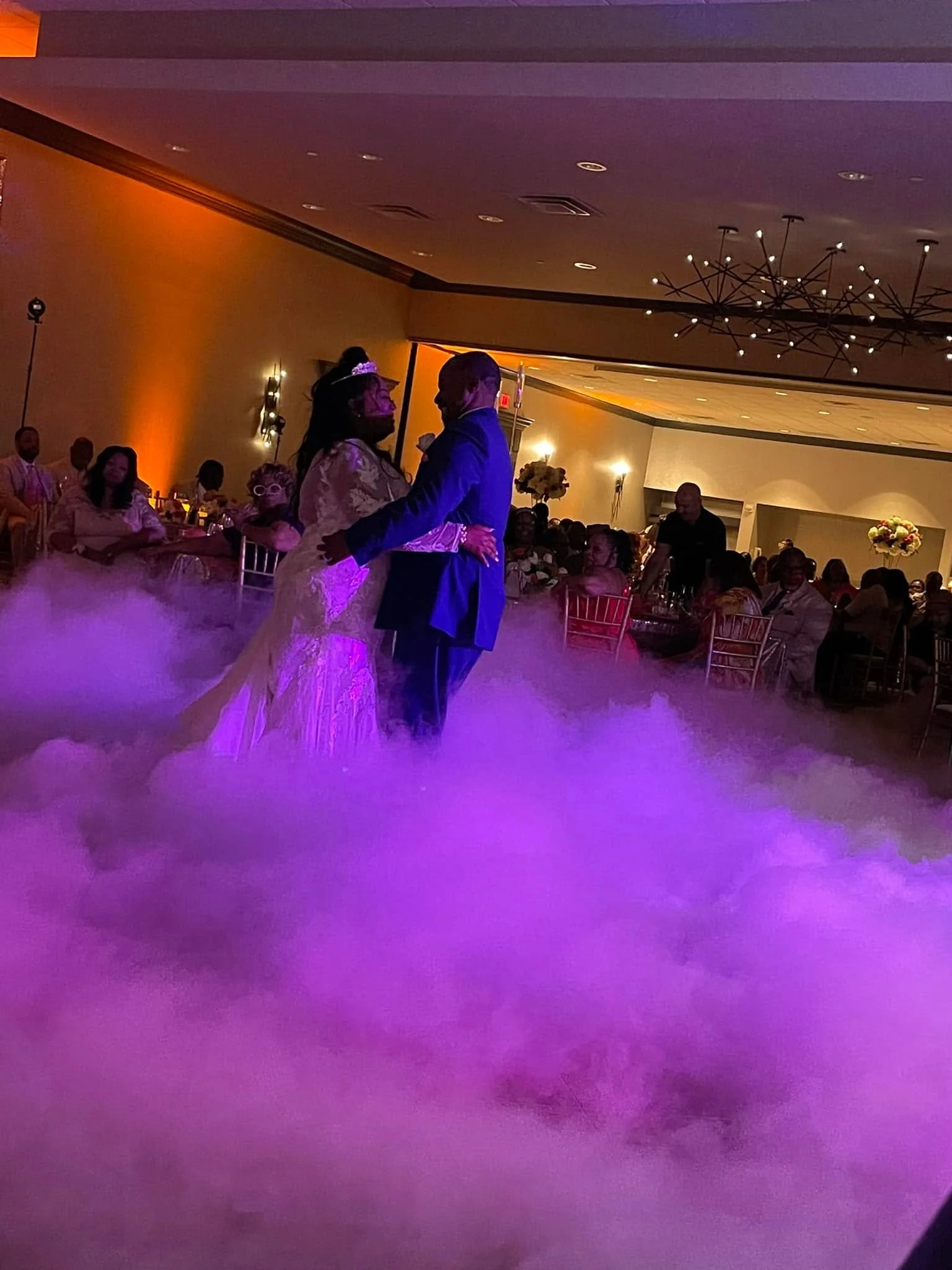 Bride and groom dancing on low0lying fog at their wedding reception, with warm lighting and seated guests in the background.