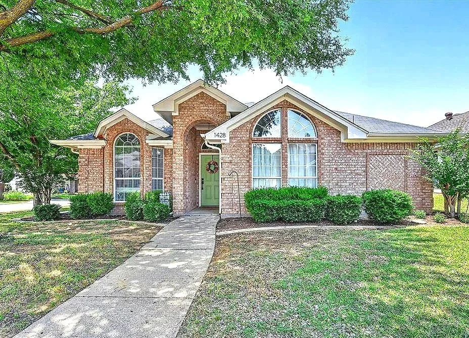 A brick house with a curved walkway leading to a green front door decorated with a holiday wreath. The house has large arched windows and well-maintained shrubbery in the front yard.