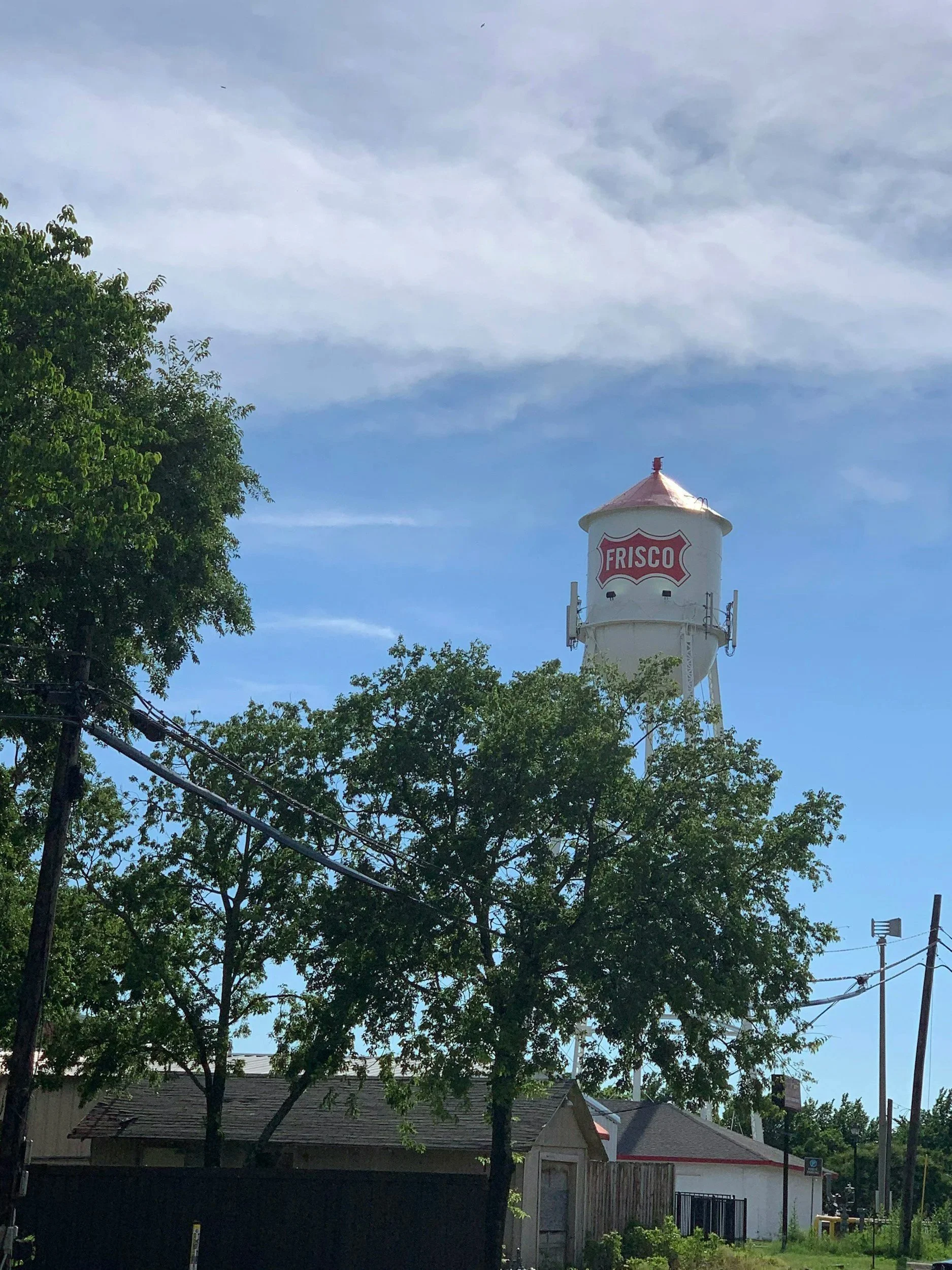 A water tower with the word 'FRISCO' written on it, framed by trees and residential houses under a cloudy blue sky.