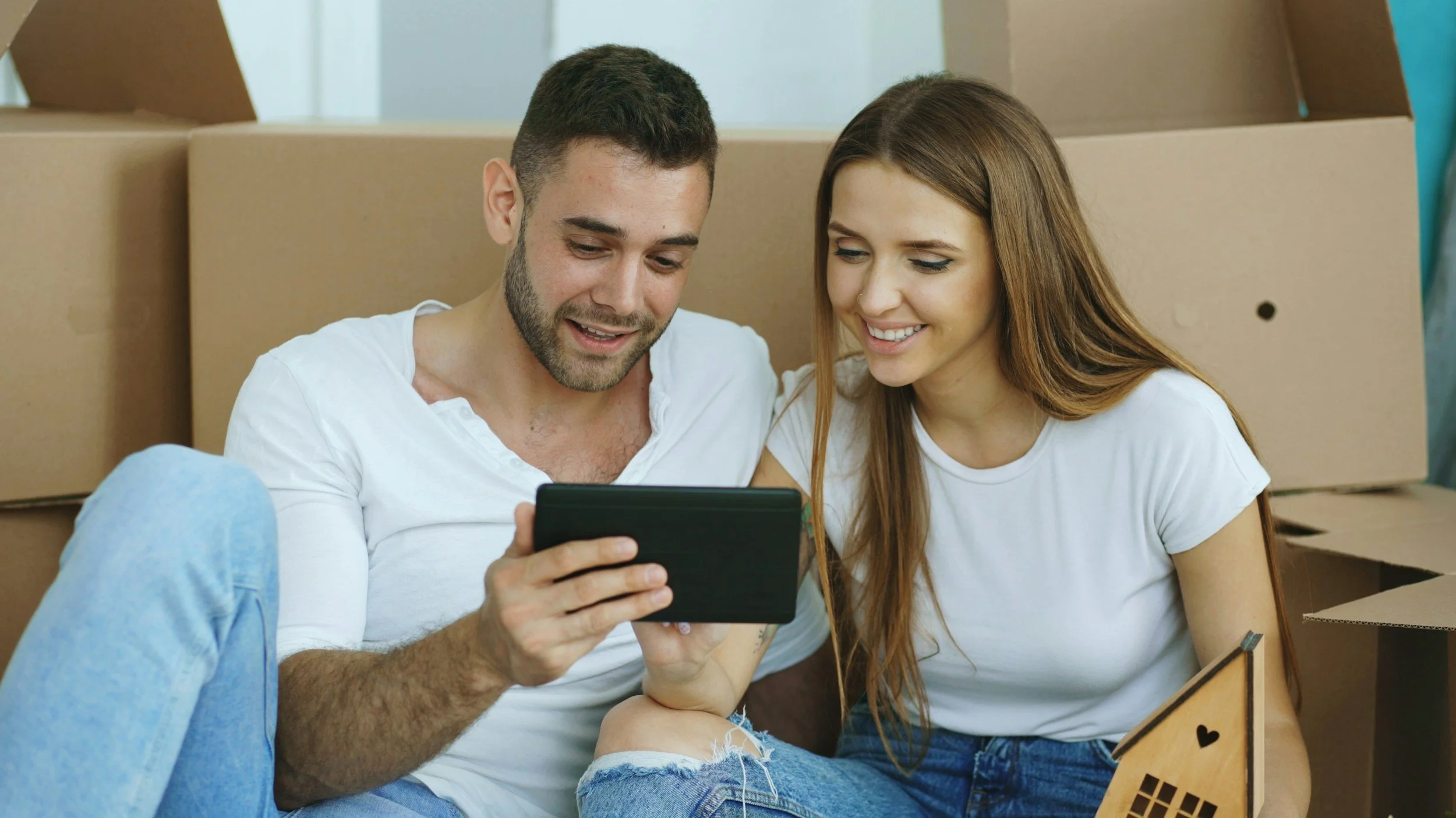 A young man and woman sit on the floor surrounded by moving boxes, looking at a smartphone together and smiling.