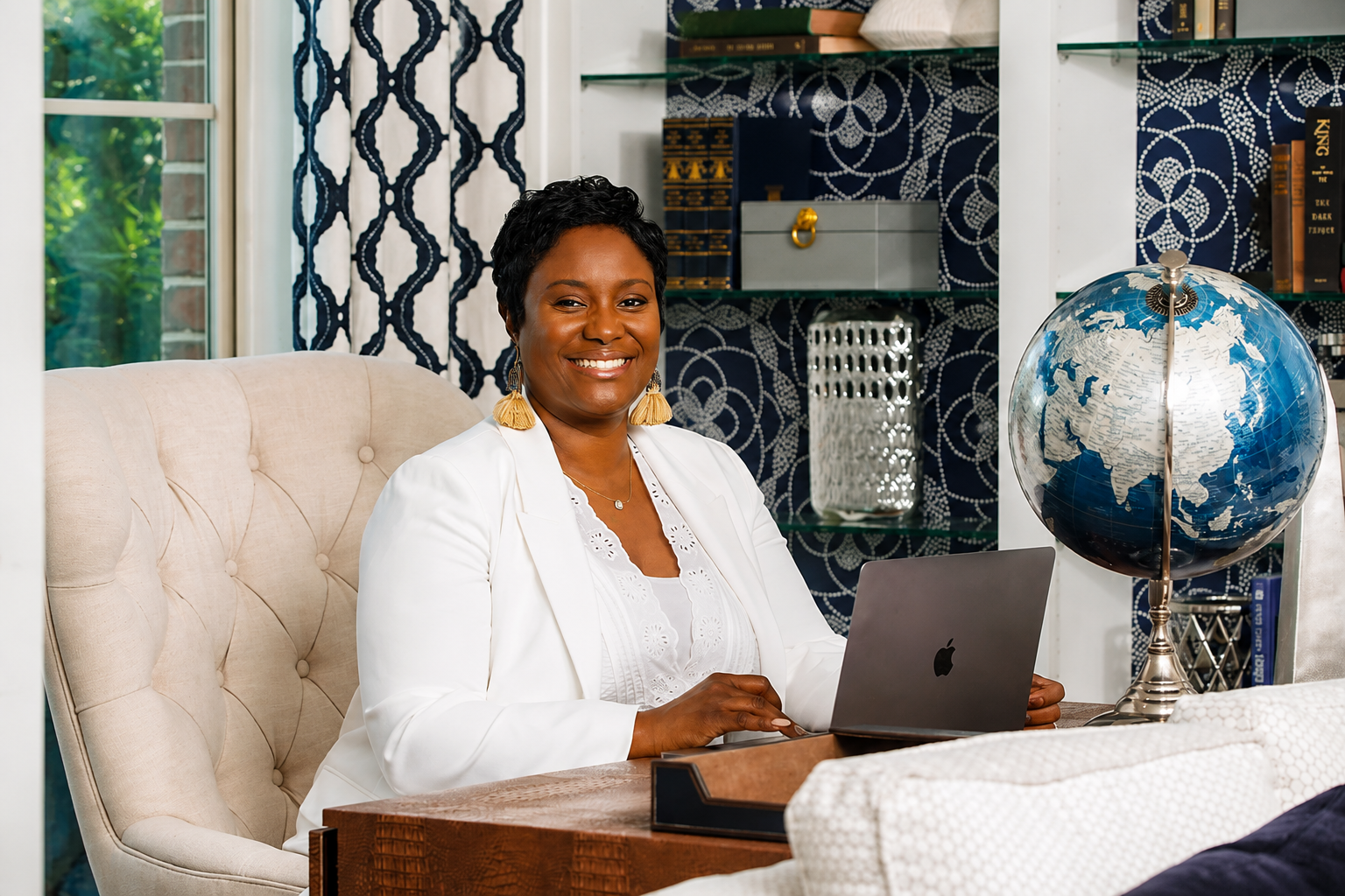 A woman with short curly black hair, wearing a white blazer, yellow tassel earrings, and a white blouse, sitting at a wooden desk with an open laptop, smiling in a stylish home office with a globe on the desk and patterned blue and white wallpaper.