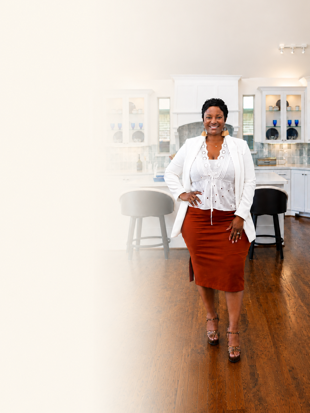 A woman smiling confidently, standing in a modern kitchen with white cabinets, a marble backsplash, and wooden flooring. She is wearing a white blazer, white top, rust-colored skirt, and high heels.