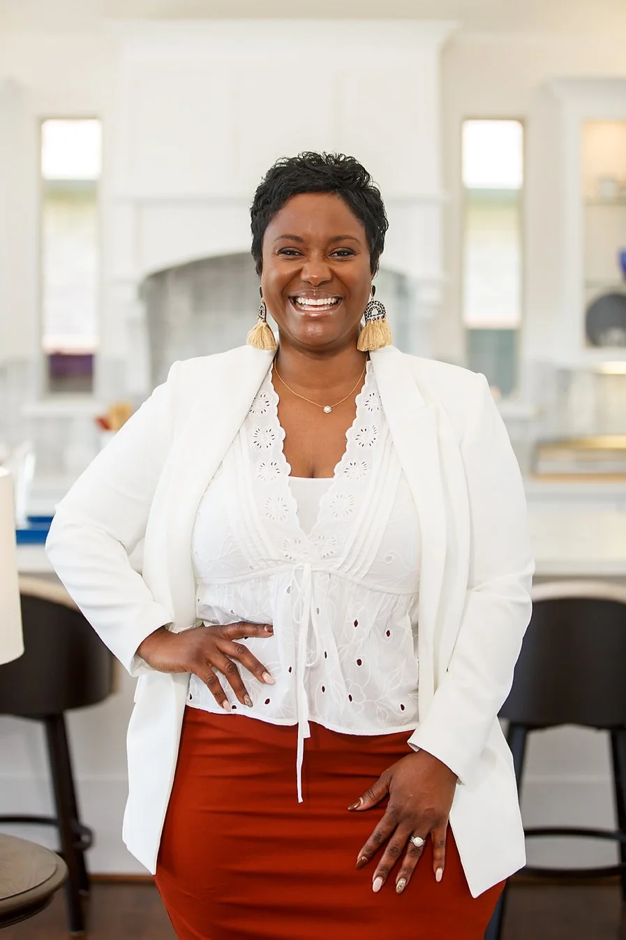 Woman smiling in a bright kitchen, wearing a white blazer, white embroidered blouse, and a rust-colored skirt.