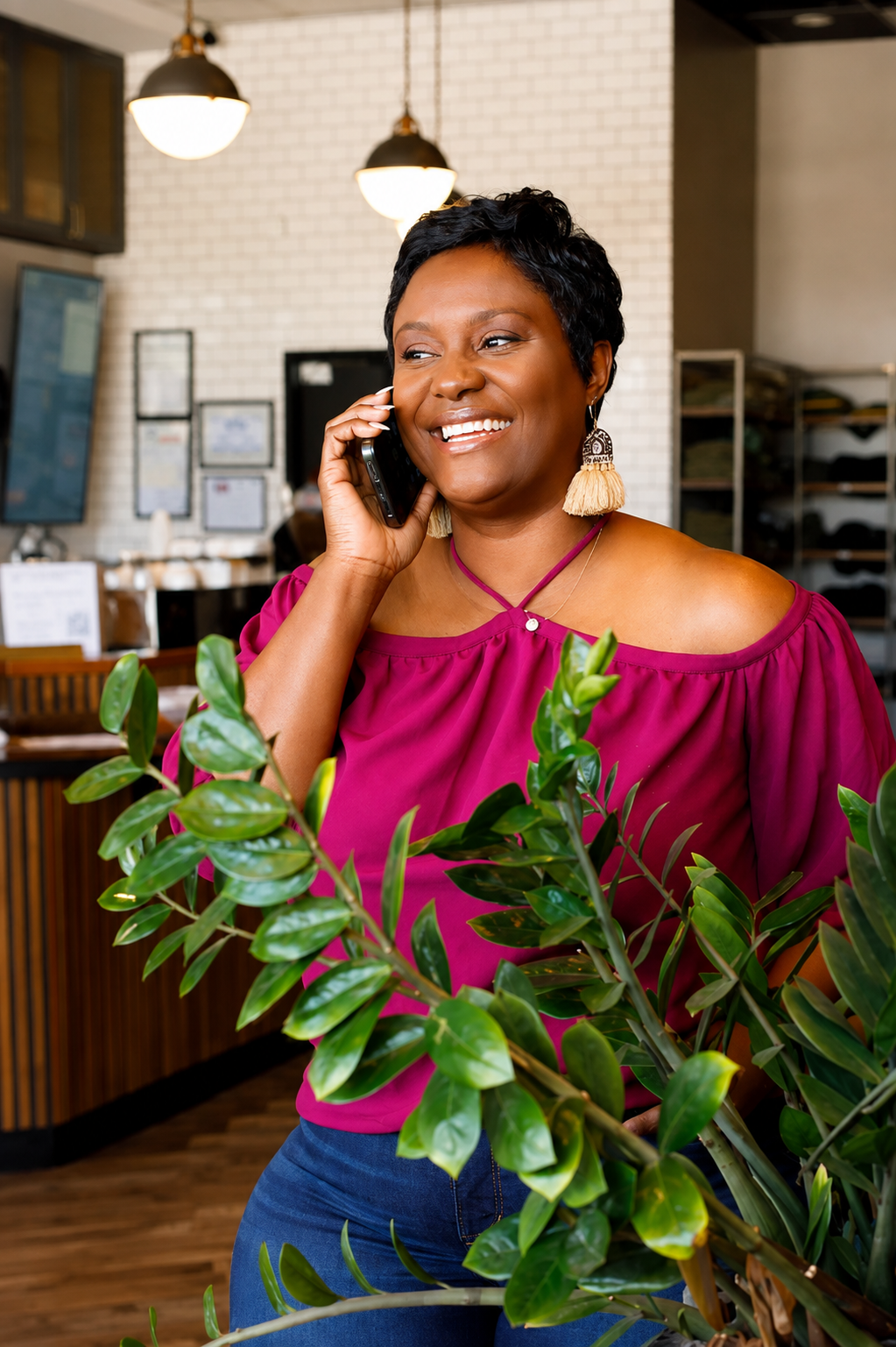 A woman with short black hair, wearing a magenta off-the-shoulder top and large earrings, is smiling and talking on a cell phone inside a restaurant or cafe. There are green plants in the foreground and warm lighting overhead.