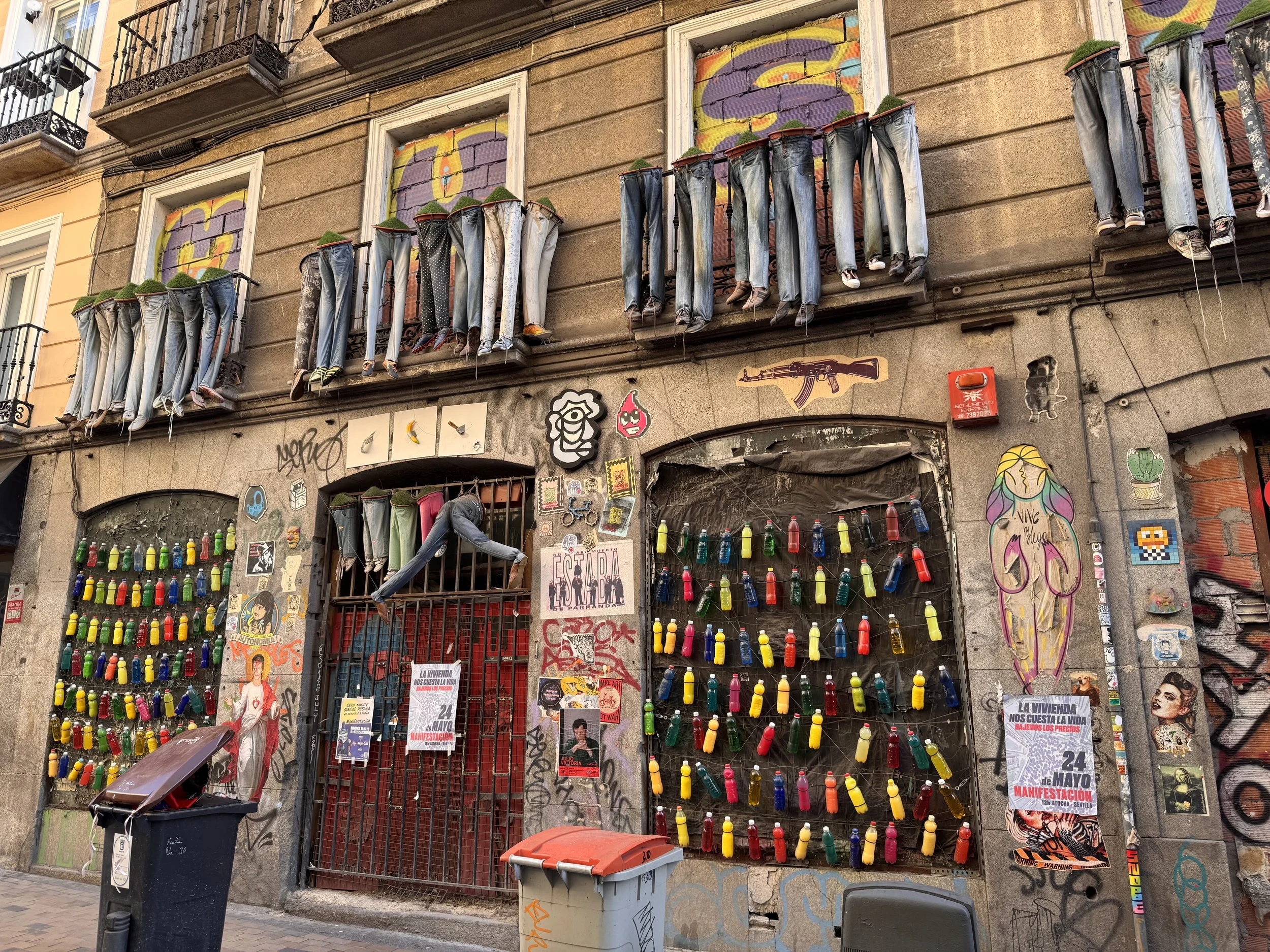 A building facade painted with colorful graffiti art, featuring three windows with numerous pairs of jeans hanging from the windowsills as planters with greenery. The ground floor has various street art, stickers, and posters, including a sign related to a May Day demonstration. There is a black trash bin and a smaller orange and white container in front of the building, with some graffiti and stickers on the walls.
