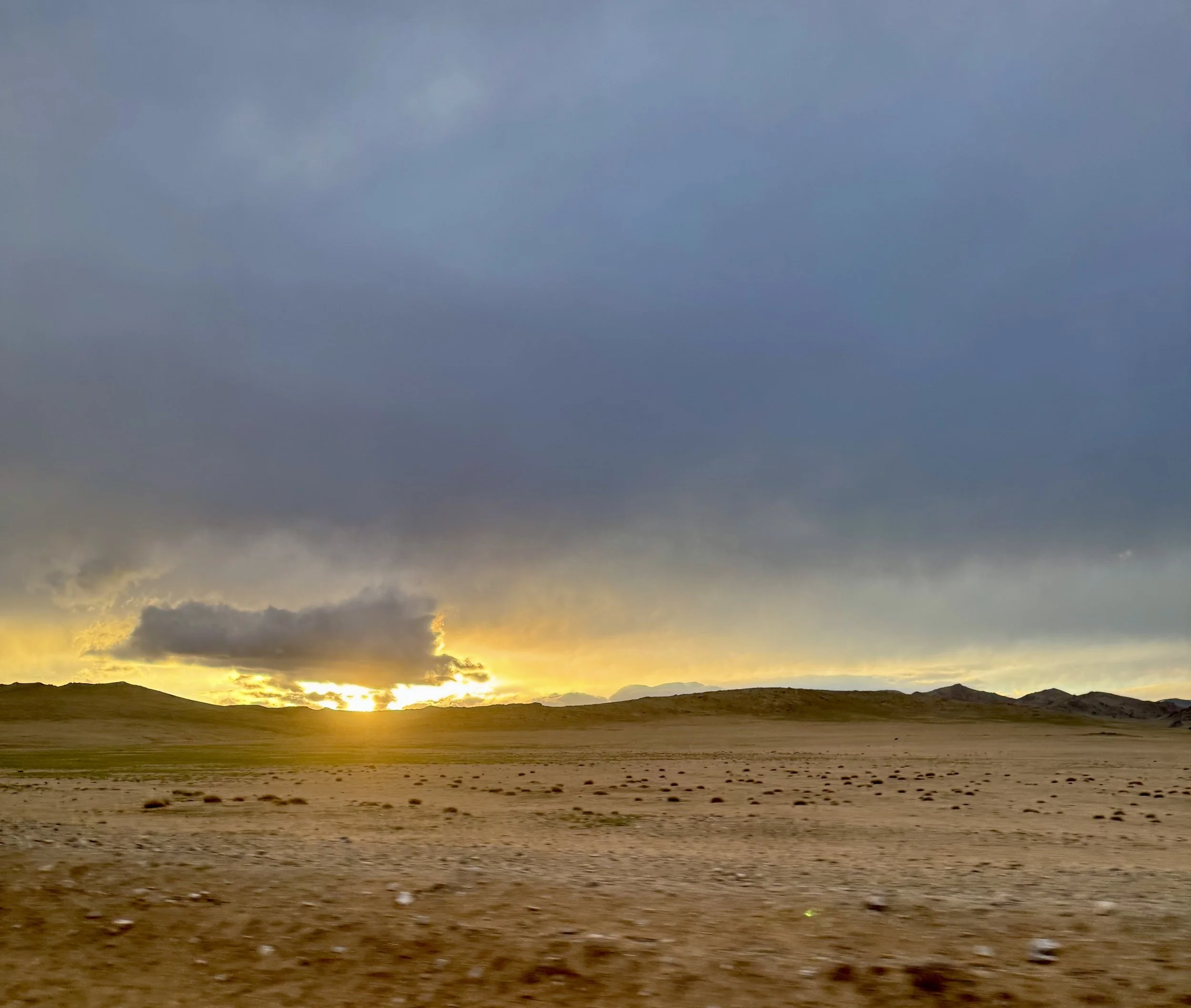 A vast desert landscape in the Eastern Cape of South Africa with rolling hills under a cloudy sky at sunset or sunrise, with the sun partially obscured by clouds on the horizon.